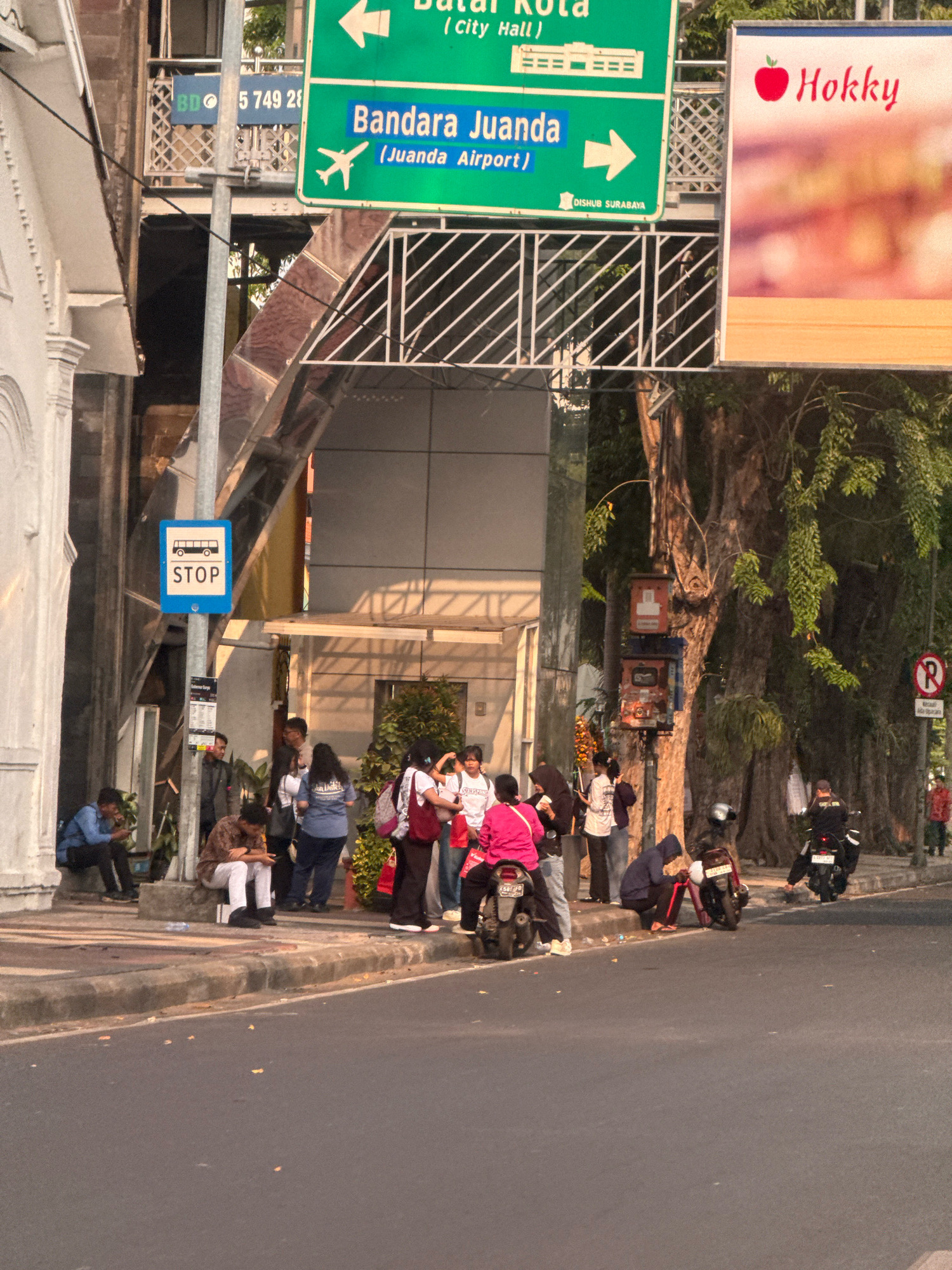 The image captures a street scene in Surabaya, Indonesia, with a bus stop sign and directional signage prominently displayed. The bus stop sign, featuring a white bus icon and the word "STOP" in blue, indicates a designated waiting area. Above it, green directional signs point towards "Batal Kota (City Hall)" and "Bandara Juanda (Juanda Airport)," with an airplane icon and an arrow indicating the airport's direction. A red apple graphic with the word "Hokky" is visible on a separate advertisement board.
Several individuals are gathered near the bus stop, with some sitting and others standing. One person in a blue shirt is seated on a concrete ledge, looking at a mobile device. Another person in a patterned shirt and white pants is also seated, looking down. A group of people, some holding bags and wearing casual attire, are congregating near the bus stop pole. Two motorcycles are parked on the street, one with a rider wearing a black jacket and helmet, and another with a rider in a black jacket and red pants. A no-parking sign with a red circle and a diagonal bar over an 'R' is visible on a pole in the background. The environment includes a sidewalk, a street, a large tree with hanging foliage, and a building with a white facade. The overall lighting suggests it is either early morning or late afternoon, with long shadows present.