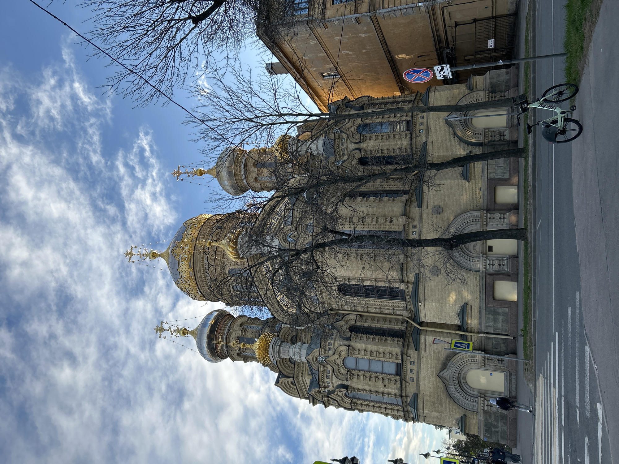 The image shows the Chesme Church in Saint Petersburg, Russia, viewed from a low angle. The church has a yellowish facade with ornate details and several domes topped with crosses. Bare tree branches partially obscure the view of the church and the blue sky with scattered clouds. A street runs alongside the church, with a pedestrian crossing visible. A bicycle is parked on a lamppost. A building is visible adjacent to the church. A pedestrian is crossing the street. A no-parking sign is present.