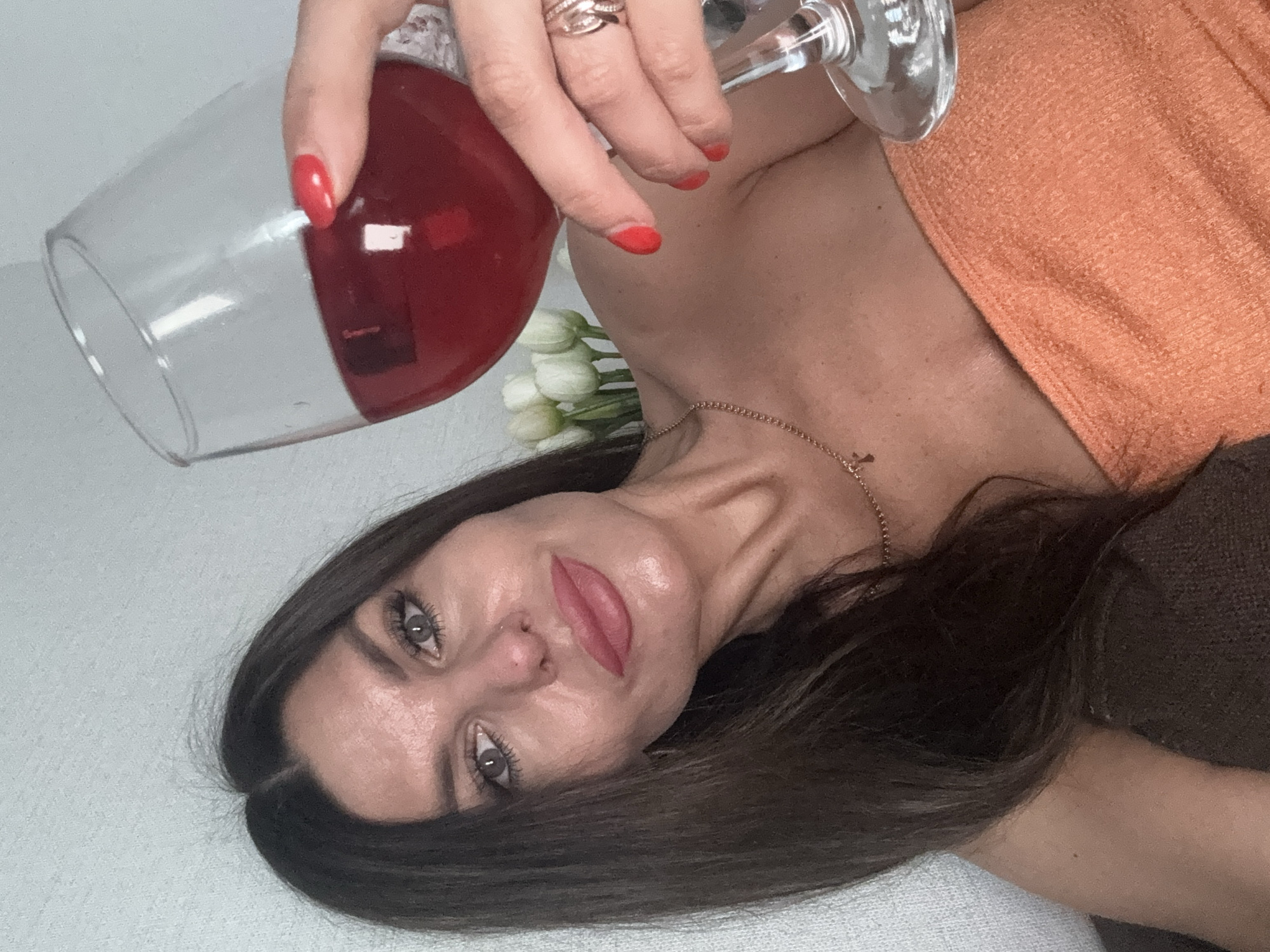 A woman is lying down, holding a glass of red wine. She has long, dark hair and is wearing makeup. Her nails are painted red. A portion of her shoulder and chest are visible, and she is wearing a gold necklace. She appears to be indoors, possibly in a bedroom or living room, as indicated by the soft lighting and the presence of what looks like bedding or a couch. There are some white flowers in the background. The image is a close-up, with the woman's face tilted towards the camera. The overall mood seems relaxed and intimate. There is no visible text in the image.