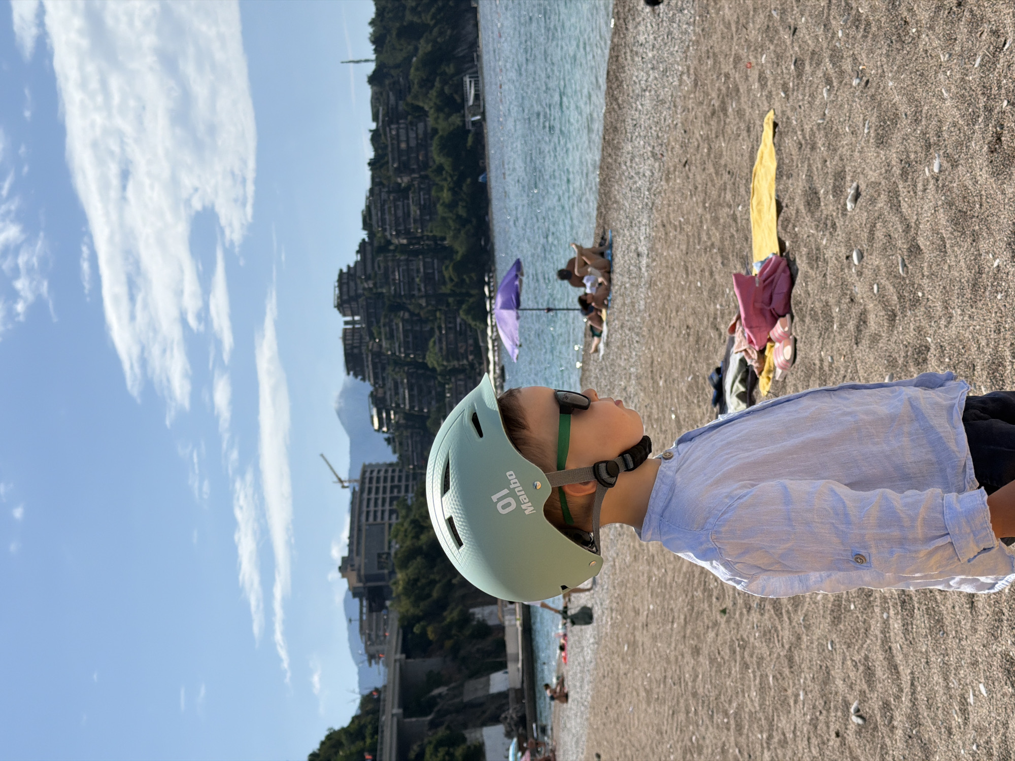 A child wearing a light blue helmet is standing on a beach. The beach is pebble-covered. In the background, there is the sea and an urban development on a hillside. The location is Boreti, Montenegro. There is a purple umbrella and several people in the background. Some personal items, including what appear to be clothes and a blanket, are on the beach nearby.