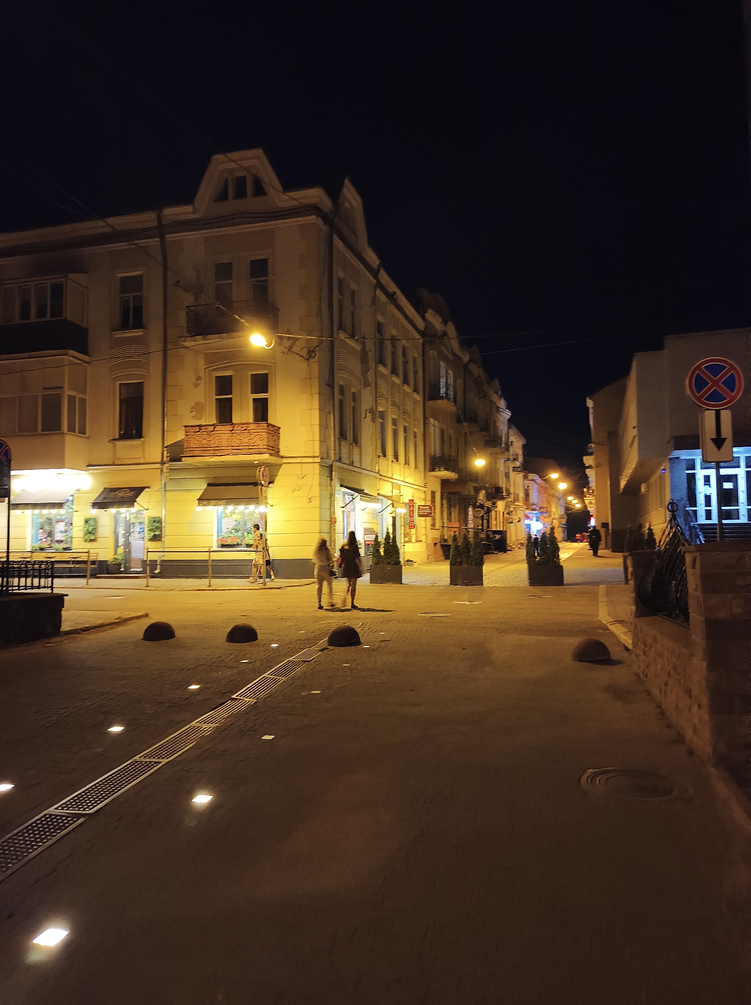 The image depicts a street scene at night in Ternopil, Ukraine, likely in a commercial or pedestrian area. A multi-story building with illuminated windows and balconies occupies the left side of the frame. On the right, another building features a blue entrance with lights and a distinctive traffic sign with a red circle and a blue cross, indicating no stopping or parking, accompanied by a downward-pointing arrow. The street is paved with interlocking bricks and features several decorative planters containing small evergreen trees. Bollards, possibly to prevent vehicle access, are visible along the sidewalk, as are small, embedded lights. A metal grating runs along the middle of the pedestrian area. Three individuals are walking on the street: two women are closer to the foreground, while a third person is further down the street, walking away from the camera. The lighting is predominantly from streetlights and building interiors, casting warm yellow and white glows on the scene. The sky is dark, indicating nighttime.