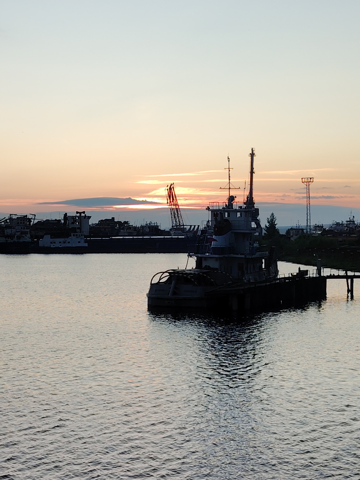 The image depicts a twilight scene at a port in Chaikovsky, Russia, with several ships and industrial structures silhouetted against a colorful sky. In the foreground, a dark, weathered tugboat is moored to a pier on the right side of the frame. The tugboat has a prominent wheelhouse and a low profile. In the background, a line of various ships and barges are docked, their forms indistinct due to the low light. A large construction crane with red accents is visible among the vessels, suggesting ongoing or past industrial activity. A tall utility tower with lights stands further in the background on the right. The sky displays a gradient of soft blues and oranges, with wispy clouds catching the last rays of the setting sun. The water in the foreground is calm, reflecting the muted colors of the sky and the dark shapes of the boats. The overall atmosphere is one of quiet industrial dormancy as day transitions into night.