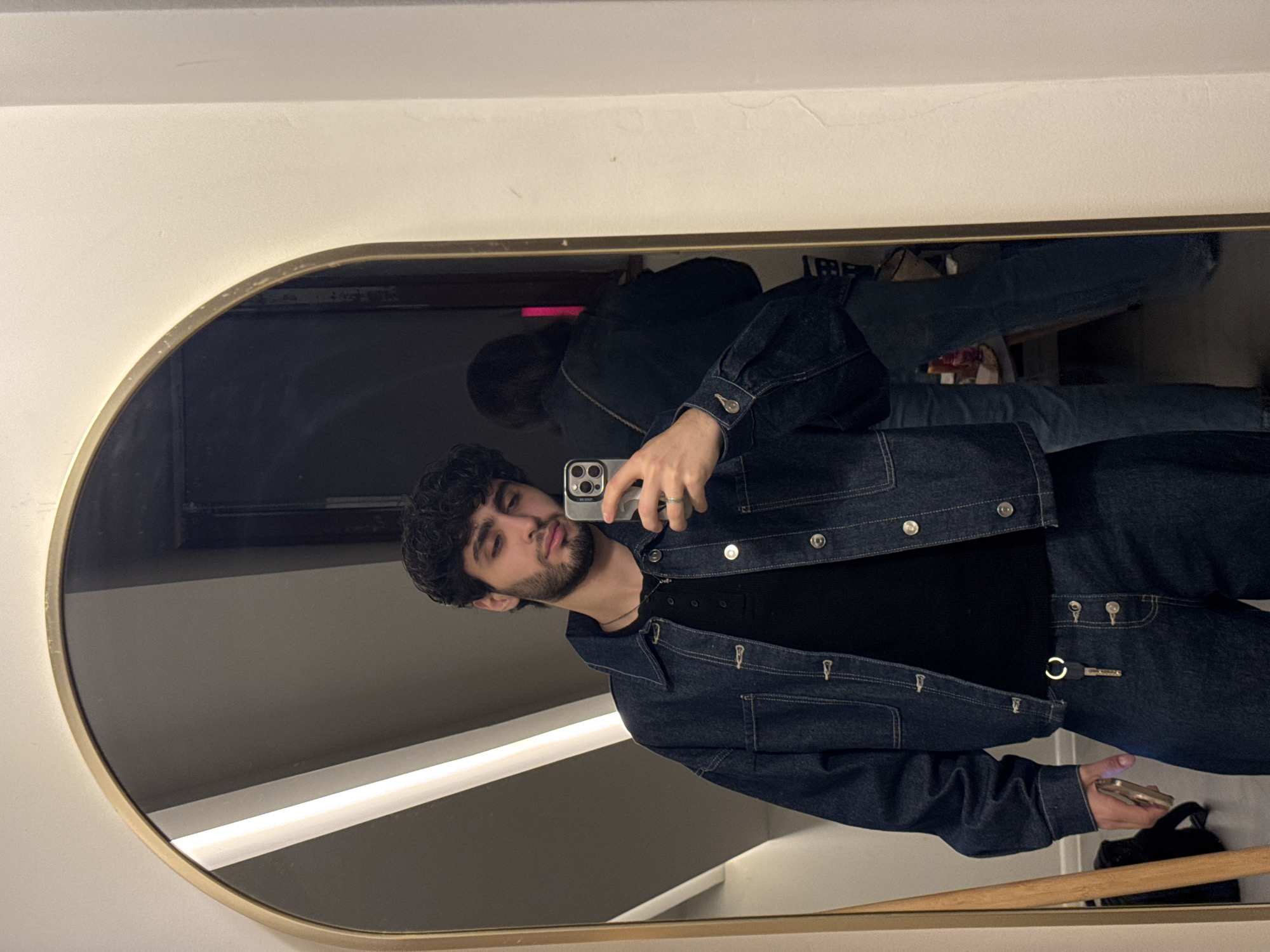 The original image is presented rotated 90 degrees counter-clockwise. It shows a mirror selfie of a young man taken indoors in Yerevan, Armenia. The man, positioned slightly to the left, is seen from the chest up, reflected in an oval or rounded rectangular mirror with a gold-colored frame. He has dark, curly hair, a full beard, and a mustache, and is looking towards the smartphone he holds in his right hand. He is wearing a dark wash denim jacket over a black t-shirt or Henley-style top, and matching dark denim pants are visible. A silver ring is on his right hand.
Behind him, in the reflection, another person or part of a person in dark clothing is partially visible. The background reflected in the mirror is dark, suggesting a dimly lit interior, possibly a hallway or lounge area, with a horizontal strip of white artificial light visible. Towards the bottom right of the mirror's reflection, a wooden handrail or bar is seen. The overall impression is a casual, indoor setting, likely in the evening or night, given the reliance on artificial lighting. No discernible text is visible in the image.