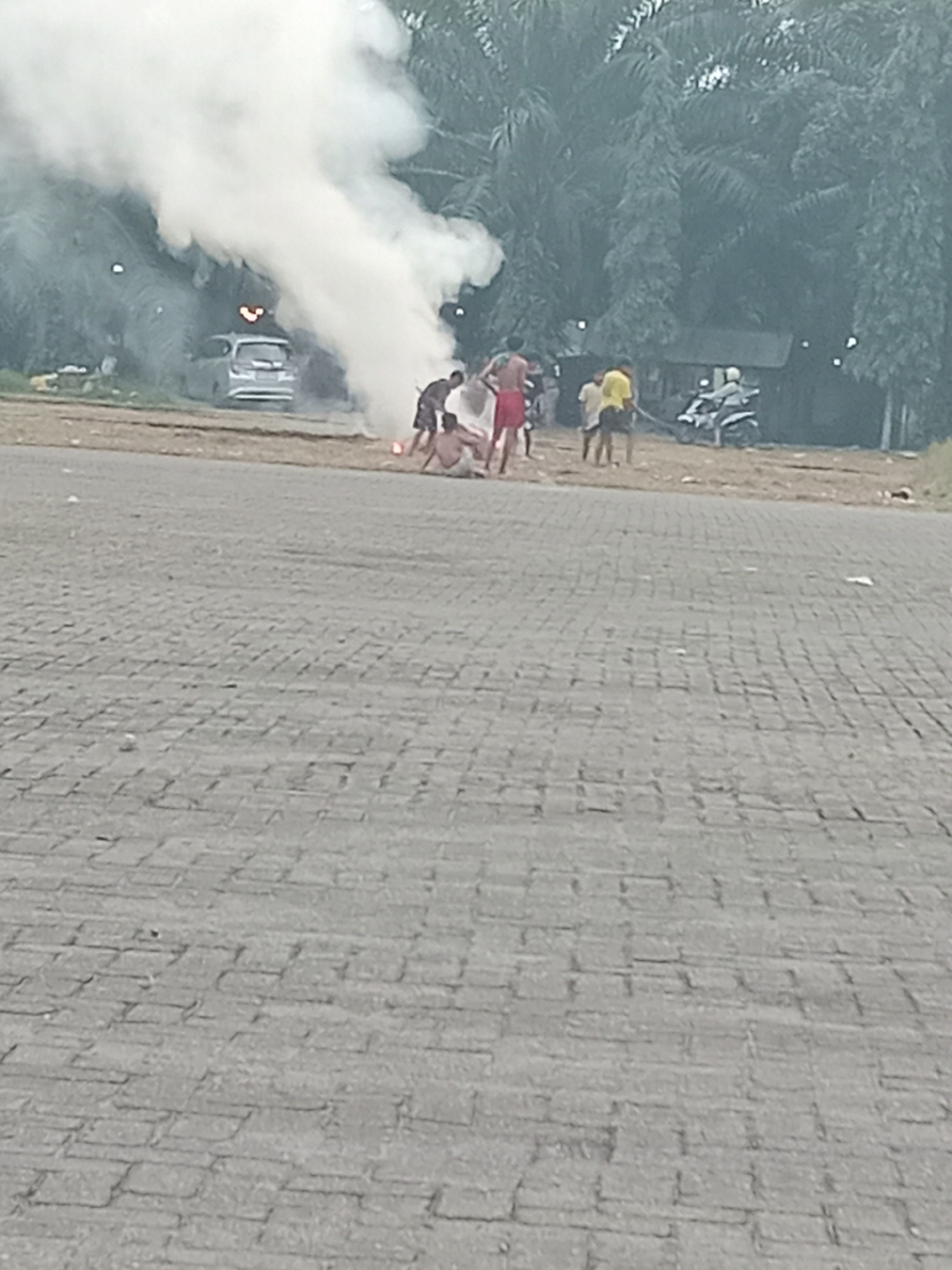 In City of Binjai, Indonesia, a group of individuals is gathered near a silver car obscured by a thick plume of white smoke. The smoke appears to emanate from the ground where some of the individuals are crouched, possibly near a fire or firework. Among the group, one person is wearing red shorts, another a yellow shirt, and others are in casual attire. In the background, palm trees and other foliage form a dense backdrop, with a small structure visible behind them. A motorcycle with a rider is also present in the scene. The ground in the foreground is paved with textured grey stones.