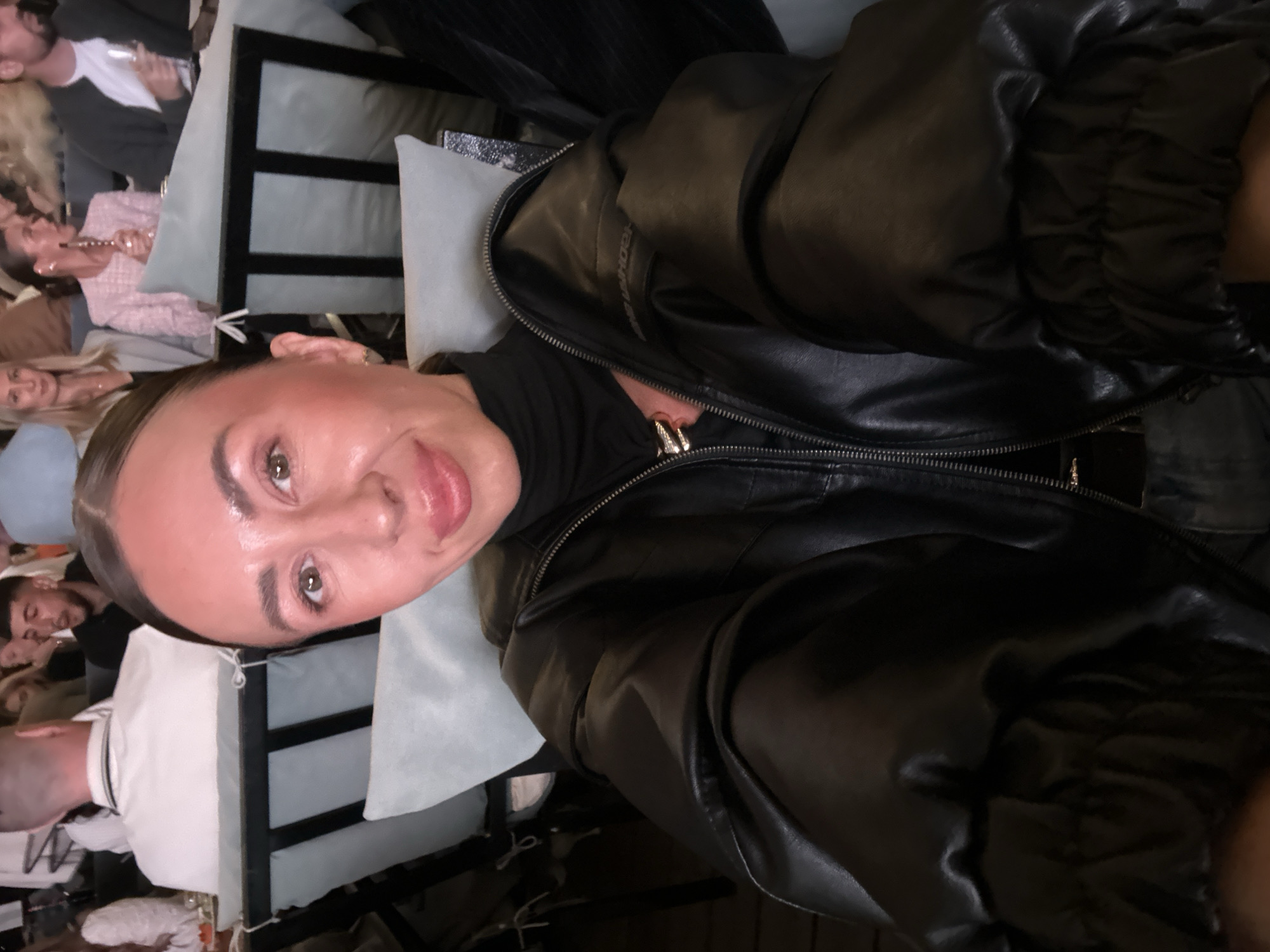 A woman with light brown hair, wearing a black turtleneck and a black leather jacket, poses for a selfie in an indoor setting. She has makeup on and is smiling. In the background, several other people are seated at tables. Some appear to be dining or drinking. The environment looks like a restaurant or a social gathering. The specific location in Izhevsk, Russia, cannot be determined from the visual information provided in the image.