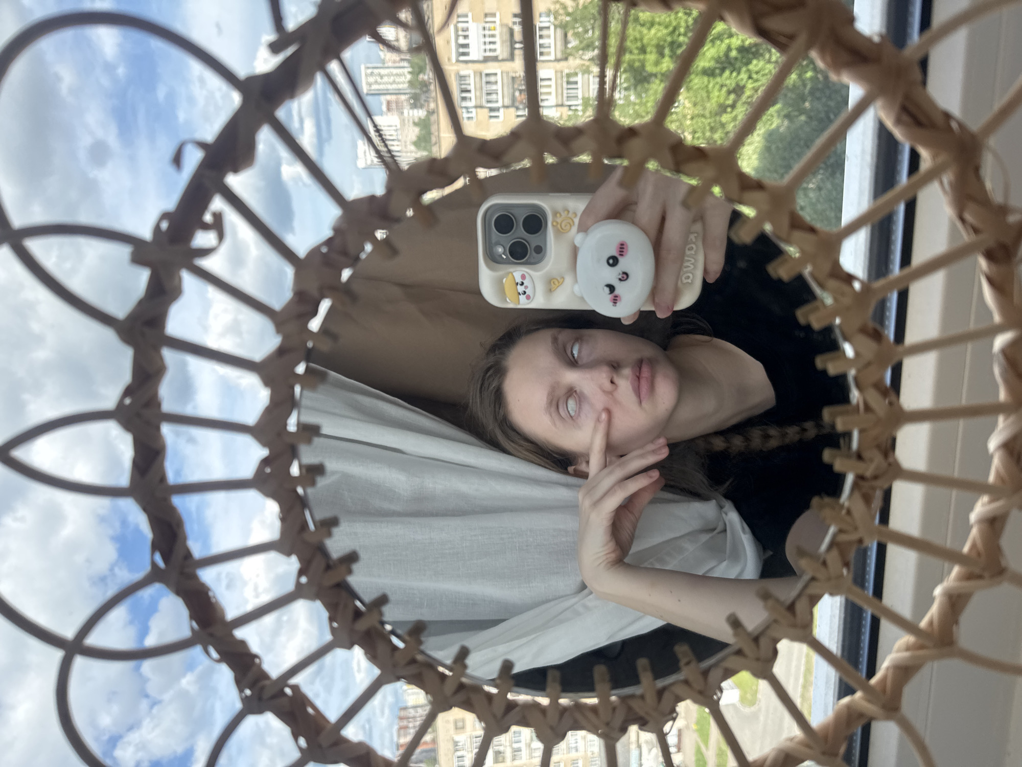 This media file is a mirror selfie showing a young person, likely in Izhevsk, Russia, captured through a decorative, circular or oval rattan frame. The image is oriented with the horizon appearing vertically.
The central subject is a person with dark hair, fair skin, and light-colored eyes that are partially rolled back, showing a lot of white. Their left hand is gently touching their cheek/eye area. They are holding a white smartphone with their right hand, which is equipped with a light-colored case featuring a small cartoon animal, a paw print, and a yellow shape. A white, circular pop-socket with a smiling cartoon bear face is attached to the back of the phone. The phone case also has faintly visible text, "KAWAII", on its side. The person is wearing a dark top, with a light-colored fabric (possibly a sheet or curtain) draped around them.
The selfie is taken within a mirror, which reflects the person and the outdoor environment. Surrounding the reflection is an intricate, woven rattan or wicker frame, forming a web-like pattern. Beyond the mirror's reflection, through a window, residential buildings with multiple stories and windows are visible, indicative of an urban setting. Green foliage from trees can also be seen. The sky is partly cloudy with patches of blue visible. The overall scene suggests a daytime setting with natural light.