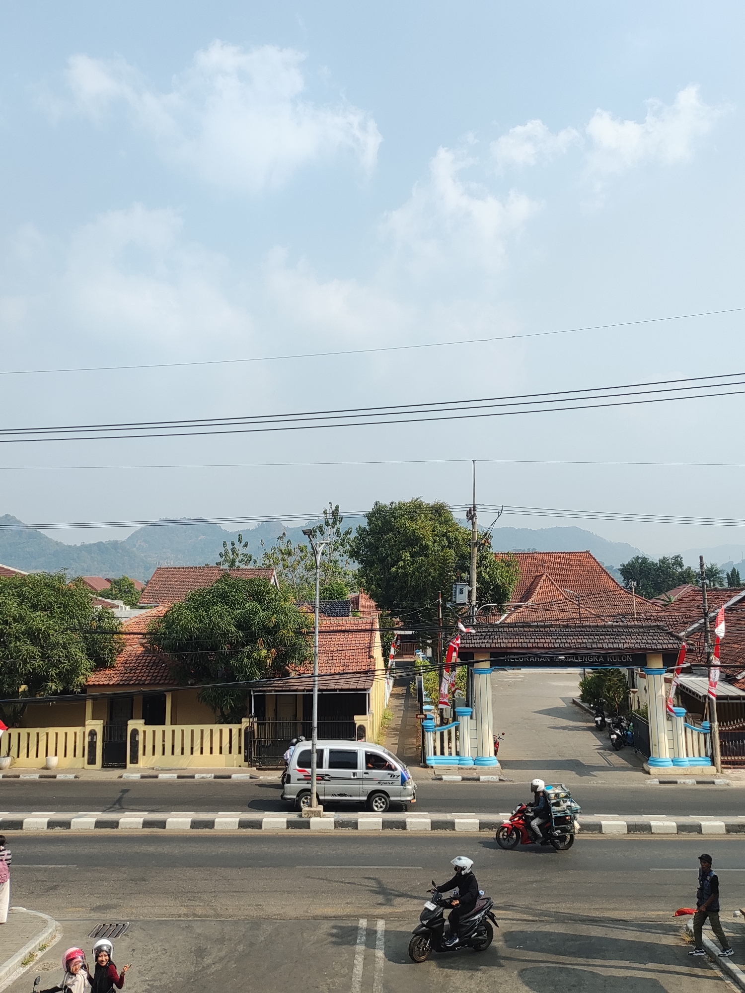 This image captures a street scene in Majalengka Kulon, Indonesia. The foreground shows a road with several vehicles and pedestrians. Two individuals wearing helmets are visible on a scooter in the lower left, and another person on a scooter is heading towards the right. A man in a vest is standing on the right, holding a red object. In the midground, a silver van is driving on the road. Behind the road, there is an arched gateway with the text "KELURAHAN MAJALENGKA KULON" inscribed on it. Beyond the gateway, houses with red tile roofs and lush green trees are visible. In the background, rolling hills are shrouded in a light haze under a pale blue sky with scattered white clouds. Power lines are strung across the upper portion of the image.