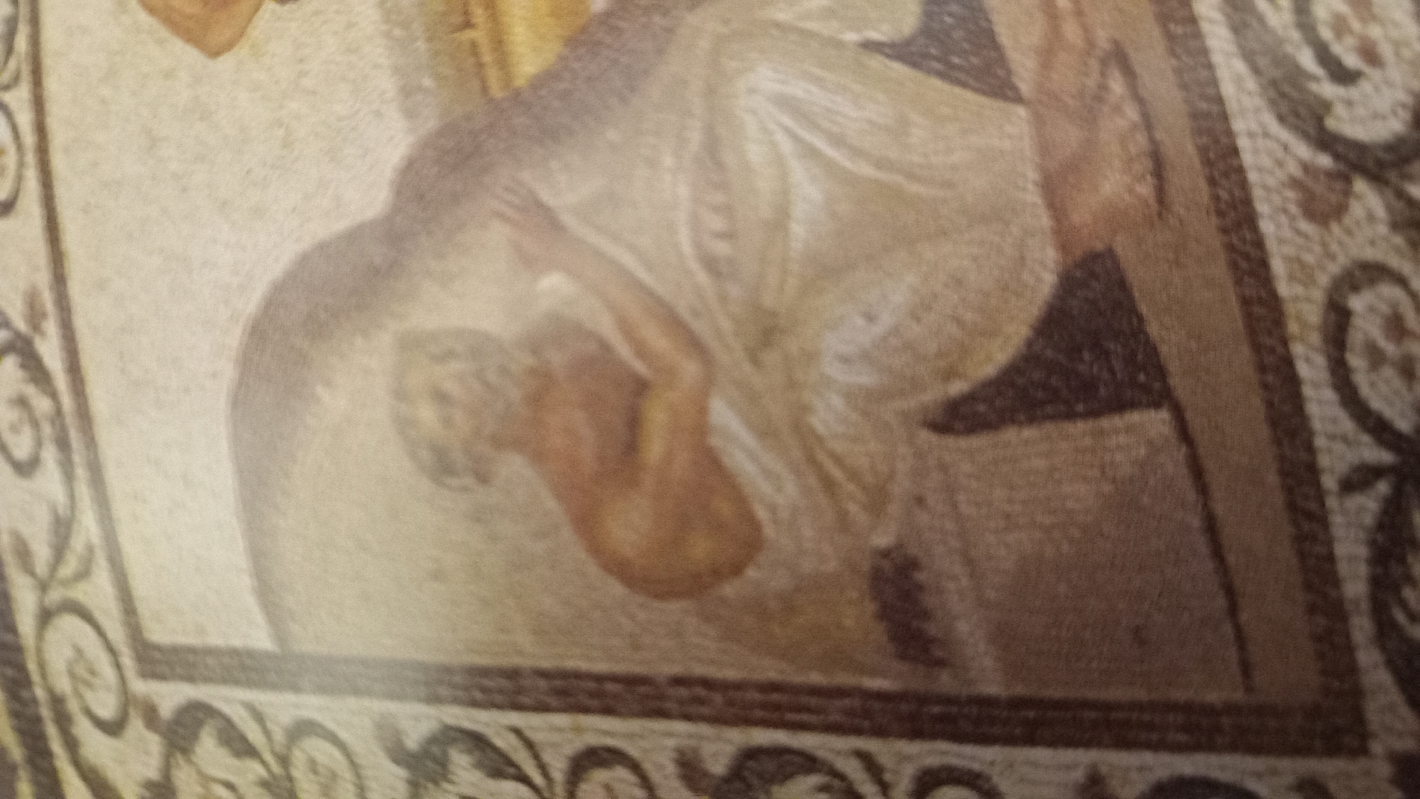 The image shows a mosaic depicting a nude figure, likely male, in a reclining position. The figure's skin tone is reddish-brown. The mosaic is framed by a decorative border with dark, scroll-like patterns. The background is a light, speckled texture, suggesting it's part of a larger mosaic. The mosaic is located in the House of Menander in the ancient city of Kos, Greece.
