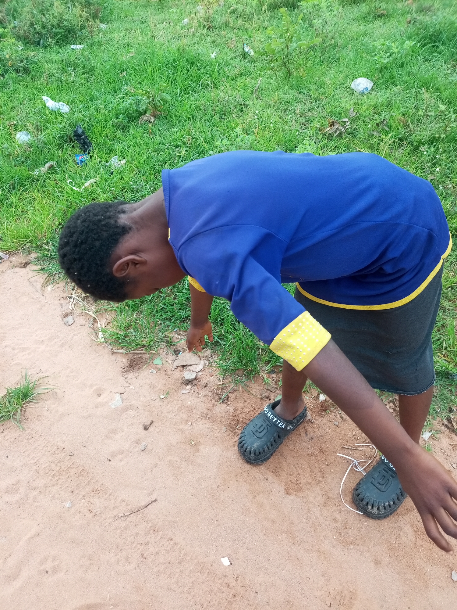 A young person, appearing to be of African descent with dark skin and black hair, is bent over in a grassy area next to a dirt path. They are wearing a blue long-sleeved shirt with yellow trim on the cuffs and neckline, and dark shorts. On their feet are black clogs with white lettering that reads "GO BETTER". The environment is a semi-urban or rural setting, characterized by green grass, dirt, and scattered litter such as plastic bags and wrappers. The person's left hand is extended towards the ground, and they appear to be examining or picking up something small. The lighting suggests it is daytime, likely outdoors. The scene does not provide specific landmarks to pinpoint a location within Tempure, Nigeria, but the general environmental conditions are consistent with such a setting.