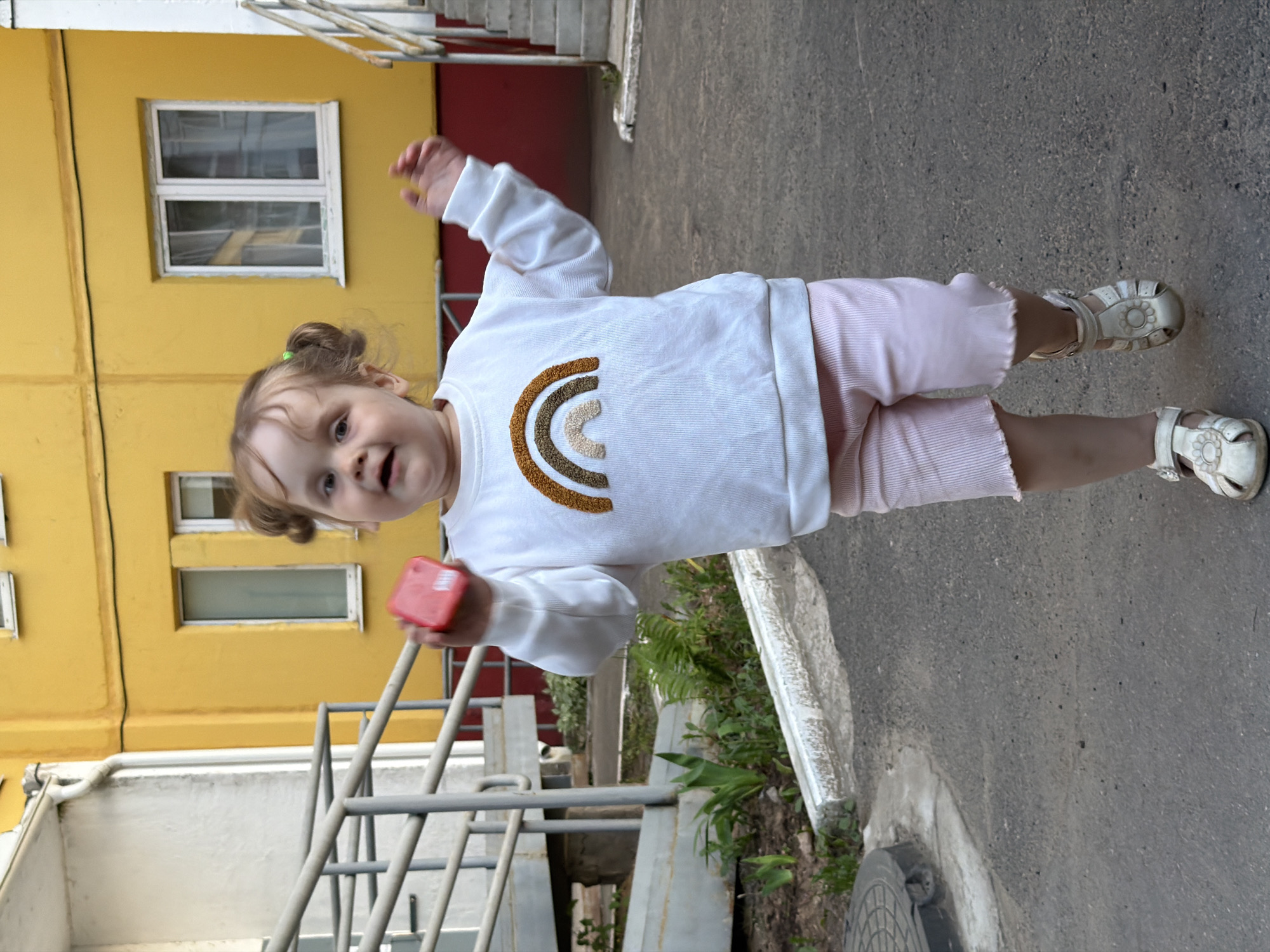 A toddler stands outdoors on a grey asphalt surface. The child wears a white long-sleeved shirt with a stitched rainbow design, light pink striped shorts with ruffled hems, and white sandals. They are holding a small red rectangular object in one hand and have their other arm raised. Their hair is styled in pigtails secured with green bands.
The background includes a yellow building with white-framed windows, a grey metal railing, and some vegetation. The setting appears to be an urban area. There is no definitive evidence that this image was taken in the city of Chaikovsky, Russia.