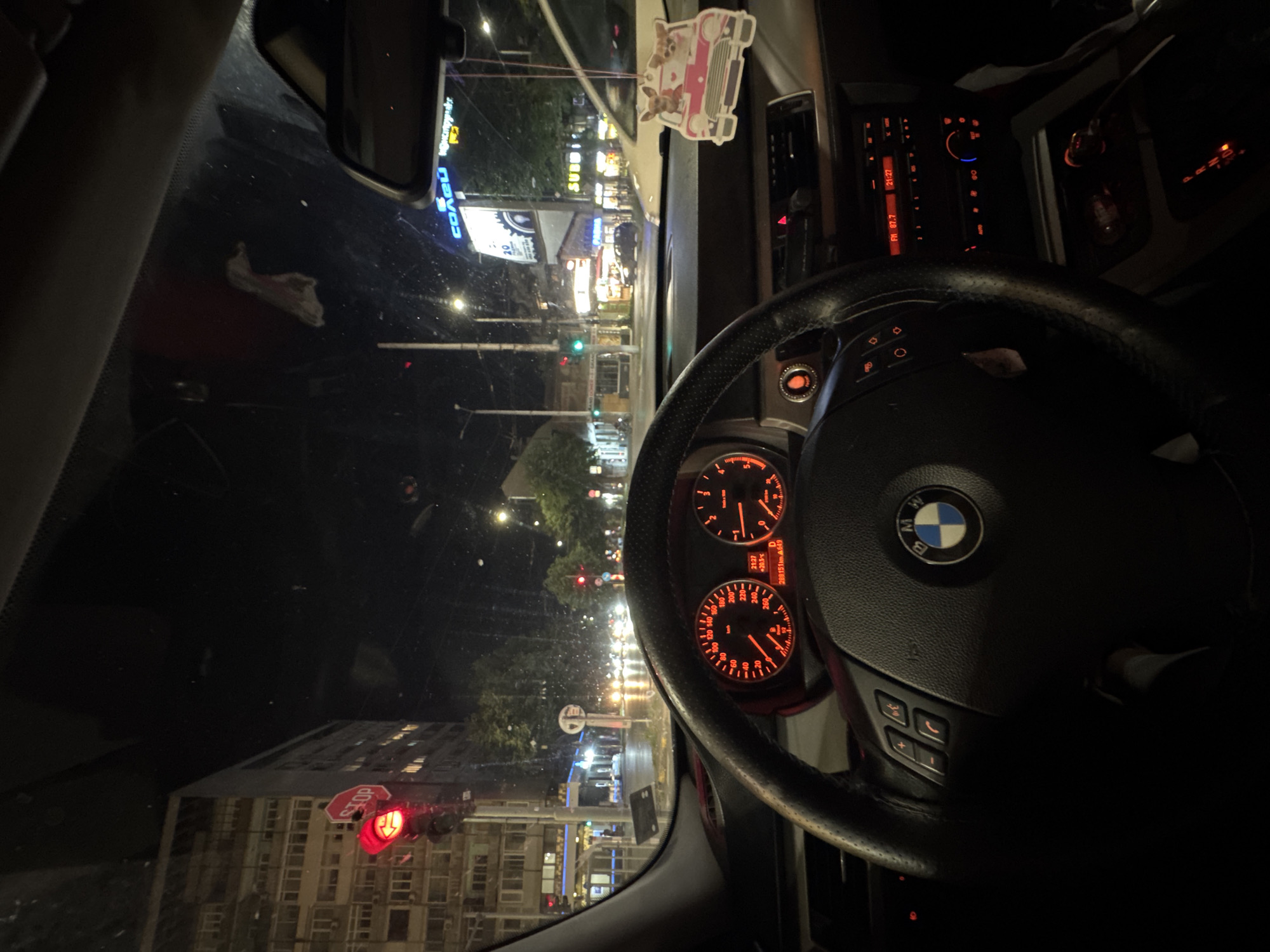 A forensic description of the image reveals a nighttime scene captured from the driver's perspective inside a BMW vehicle, specifically at the intersection of Tsar Osvoboditel Boulevard and Vasil Levski Boulevard in Varna, Bulgaria.
The vehicle's interior shows a black leather-wrapped steering wheel with multimedia and cruise control buttons. The BMW emblem is centered on the steering wheel. The instrument cluster is illuminated in red and orange, displaying a speedometer calibrated to 260 km/h and a tachometer to 7000 RPM. A digital display indicates the time as 21:27, an ambient temperature of +20.5°C, and an odometer reading of 288,151 kilometers. The gear selector display shows "P," indicating the vehicle is in park. The radio unit is also illuminated in red, showing "FM 87.7" and the time "21:27." Below the radio, climate control settings are visible. An air freshener, shaped like a pink car with two cartoon dogs, hangs from the rearview mirror. The windshield appears to have minor smudges or water marks.
Through the windshield, a city street scene unfolds at night. The road surface is wet, suggesting recent rainfall. A red traffic light is prominently visible on the left side of the frame, accompanied by a hexagonal "STOP" sign attached to its pole. In the distance, on the right side of the street, a large blue neon sign displays the Cyrillic word "СОНЦО." Below it, another illuminated sign is partially visible. Further down the street, also on the right, a yellow "SUBWAY" sign is discernible. Numerous streetlights illuminate the area. Vehicle headlights are visible in the distance, indicating oncoming or stopped traffic. A green traffic light is visible further down the road on the right. Large multi-story buildings line the street, consistent with urban architecture. No people are clearly identifiable in the visible areas of the exterior.