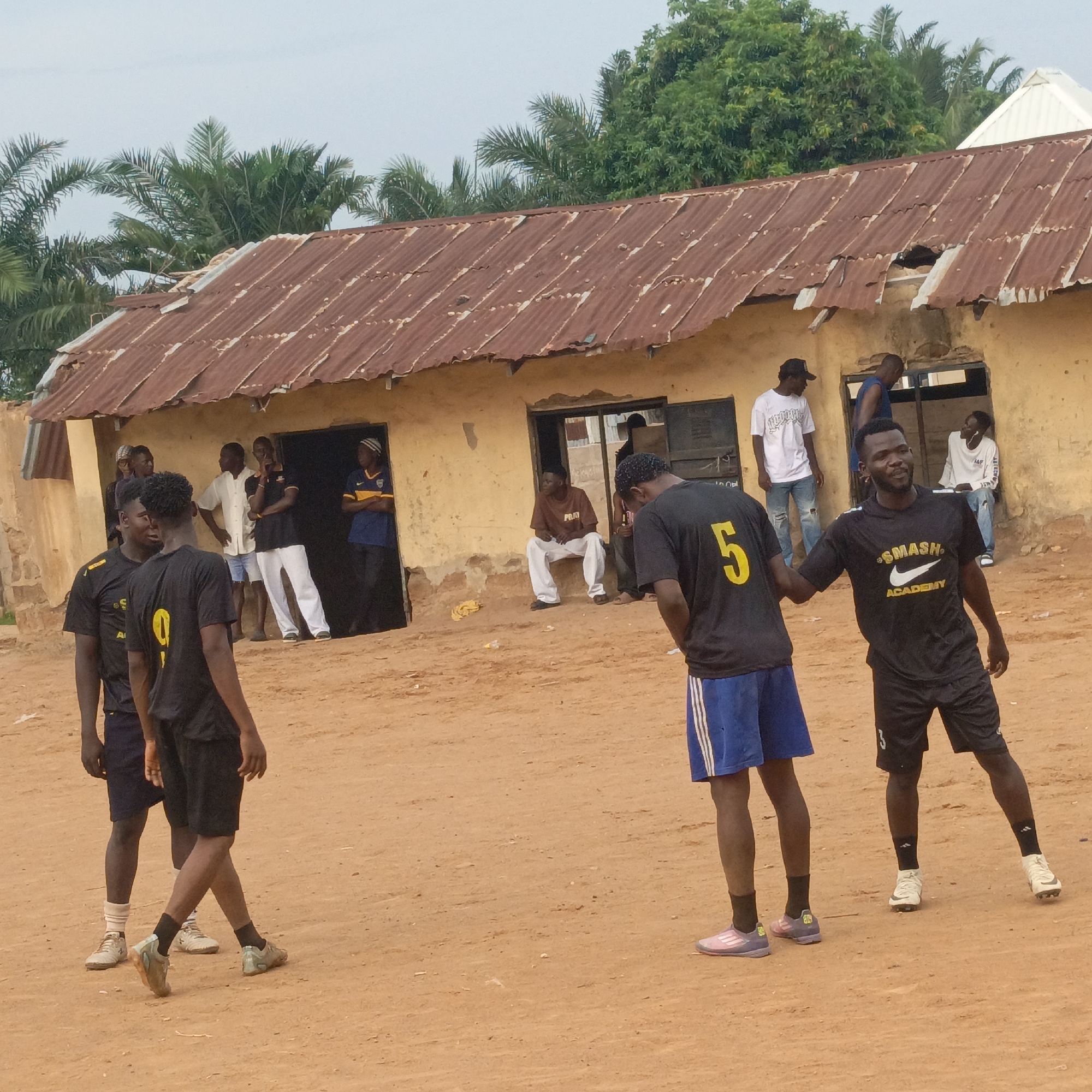 This outdoor image, taken in Akwanga, Nigeria, features a group of men gathered on a reddish-brown dirt ground in front of a rustic, light brown building with a heavily rusted corrugated metal roof. Palm trees and other green foliage are visible behind the building, set against an overcast sky.
In the foreground, several young men are in athletic attire, suggesting a sports activity. One man, facing the camera, wears a black t-shirt emblazoned with "SMASH ACADEMY" and a Nike swoosh logo in white, paired with black shorts and sneakers. He has a faint number '3' visible on his shorts. To his left, another man, seen from the back, wears a black t-shirt with a prominent yellow number '5' and blue shorts with white stripes. Another man on the far left also wears a black shirt with a yellow number '9' on the back. The footwear of some individuals appears to be athletic shoes or cleats, consistent with football (soccer).
In the background, other men are standing or sitting in and around the open doorways of the simple building. One man sits in a doorway wearing a brown t-shirt with "PMA" printed on it and white pants. Another sits nearby in a white t-shirt with "A&F" visible. The group appears to be casually socializing or taking a break from an activity. The atmosphere is relaxed, with some individuals displaying subtle smiles. The scene depicts a community gathering in a modest, possibly rural or semi-urban, setting.