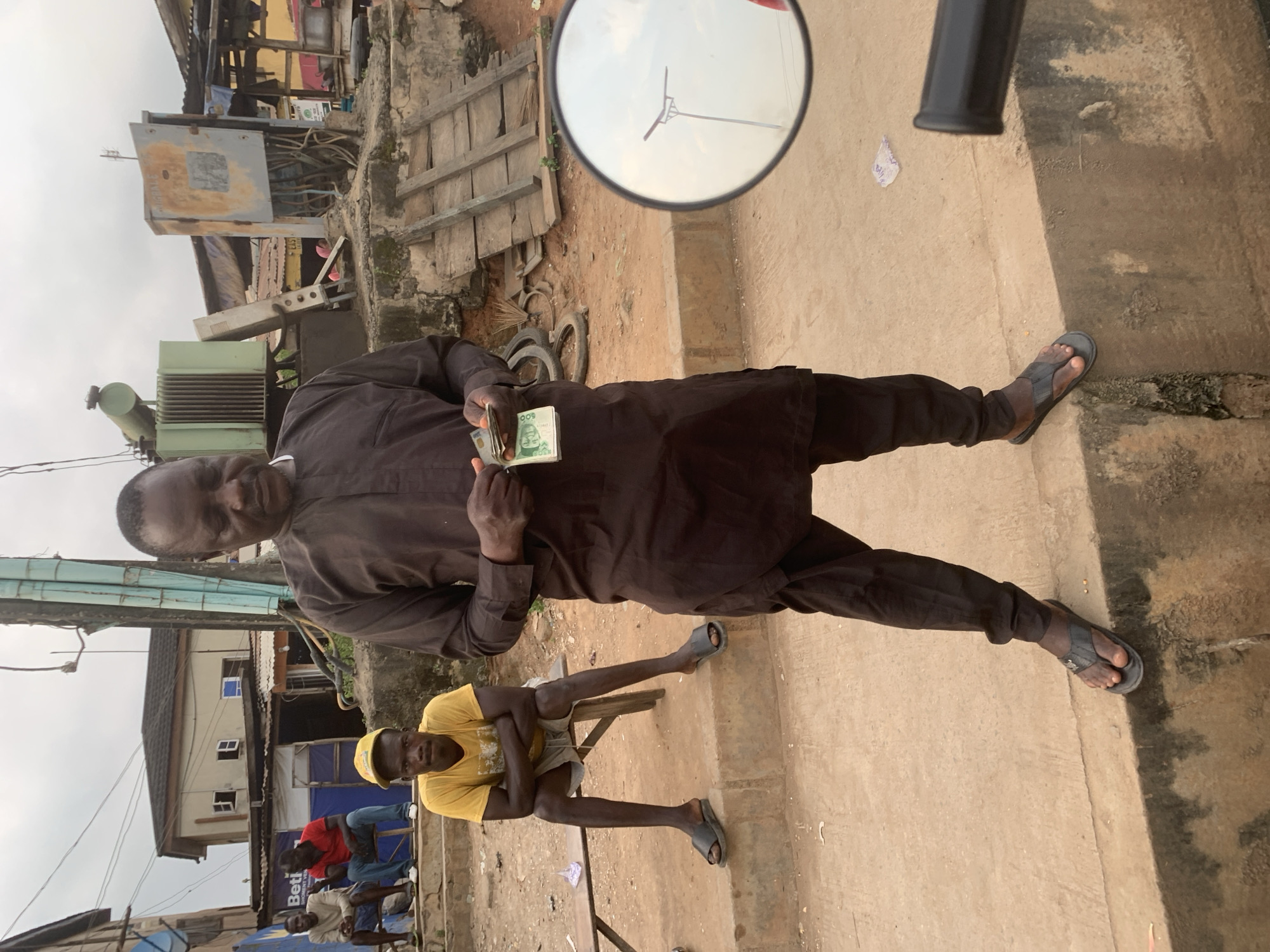 In the city of Shagamu, Nigeria, a man in a dark brown traditional outfit and flip-flops stands on a concrete ledge, holding a stack of Nigerian Naira currency. To his left, a younger man in a yellow t-shirt and shorts, wearing a yellow baseball cap and sandals, sits on a wooden bench with his arms crossed. Behind them, two other men are seated on a wooden bench; one wears a white t-shirt and blue jeans, while the other sports a red t-shirt and blue jeans. The background reveals a street scene with buildings, electrical equipment including a transformer, and makeshift stalls. The ground is paved with concrete and dirt, with debris scattered around. A motorcycle mirror is visible in the foreground, reflecting a streetlamp.
