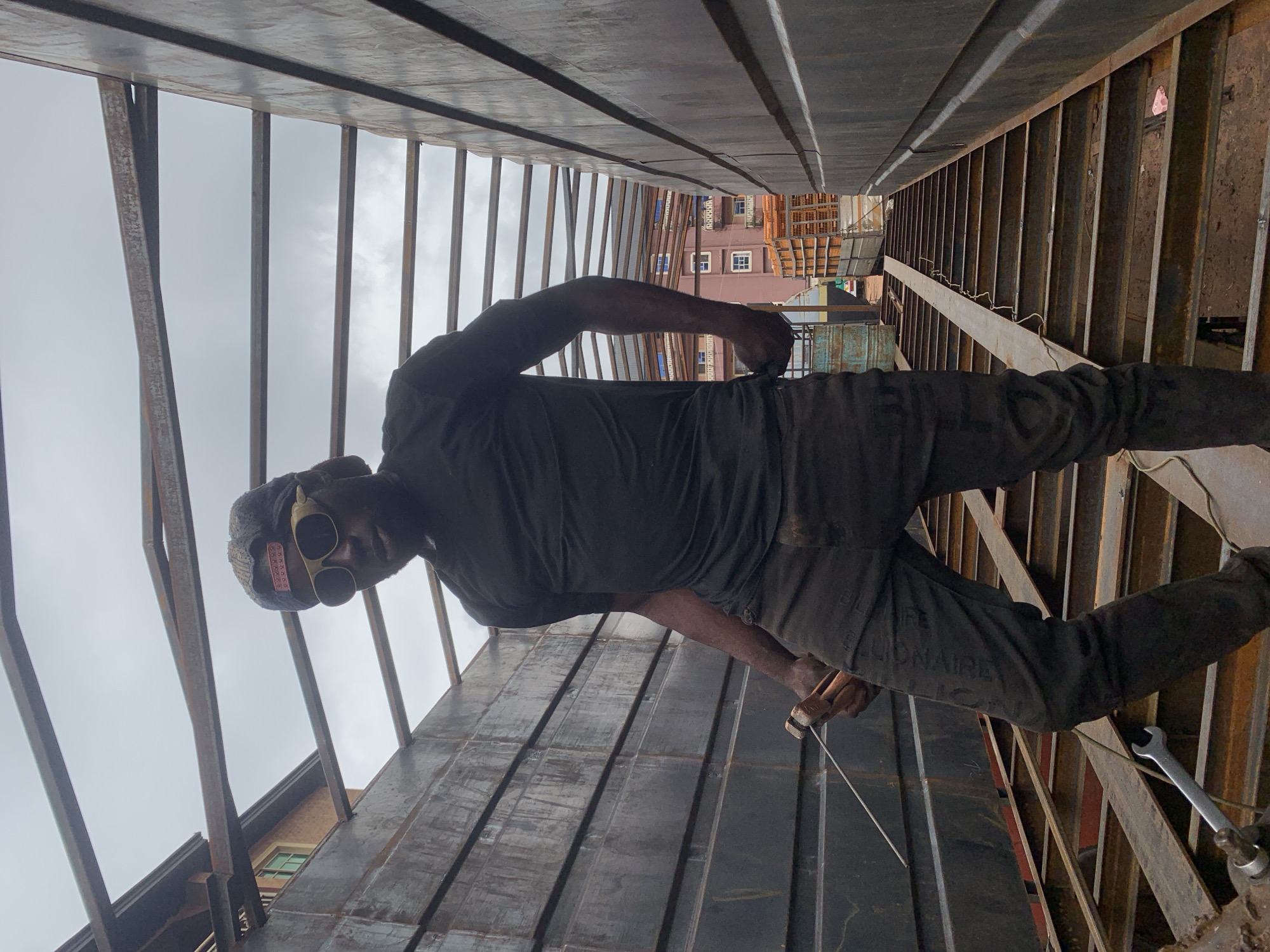 In Nkpor, Nigeria, a man wearing a black t-shirt, dark pants, a baseball cap, and gold-rimmed sunglasses is standing within a metal structure, possibly a truck bed or a workshop. He holds what appears to be a welding torch in his right hand and a thin wire in his left. The structure is made of parallel metal beams and corrugated metal walls. In the background, buildings with windows are visible, along with stacked orange crates on a truck. The sky is overcast.