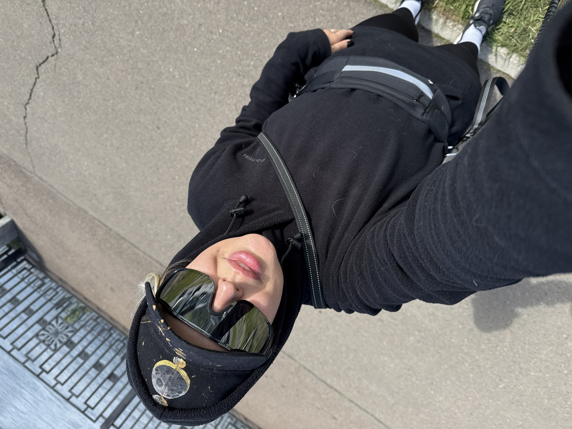 A medium-level, high-angle shot of a person lying on a gray paved surface. The person is dressed in black pants, a black hooded fleece jacket, black running shoes with white socks, a black fanny pack with a reflective stripe, and a black baseball cap with an emblem. The person is also wearing sunglasses with reflective lenses. The person's face is visible. The person appears to be taking a selfie. A decorative metal railing is partially visible at the left edge of the frame. The pavement surface has cracks. Based on this information, the image was likely taken in the city of Krasnoyarsk, Russia, near a paved area with decorative metal railing.