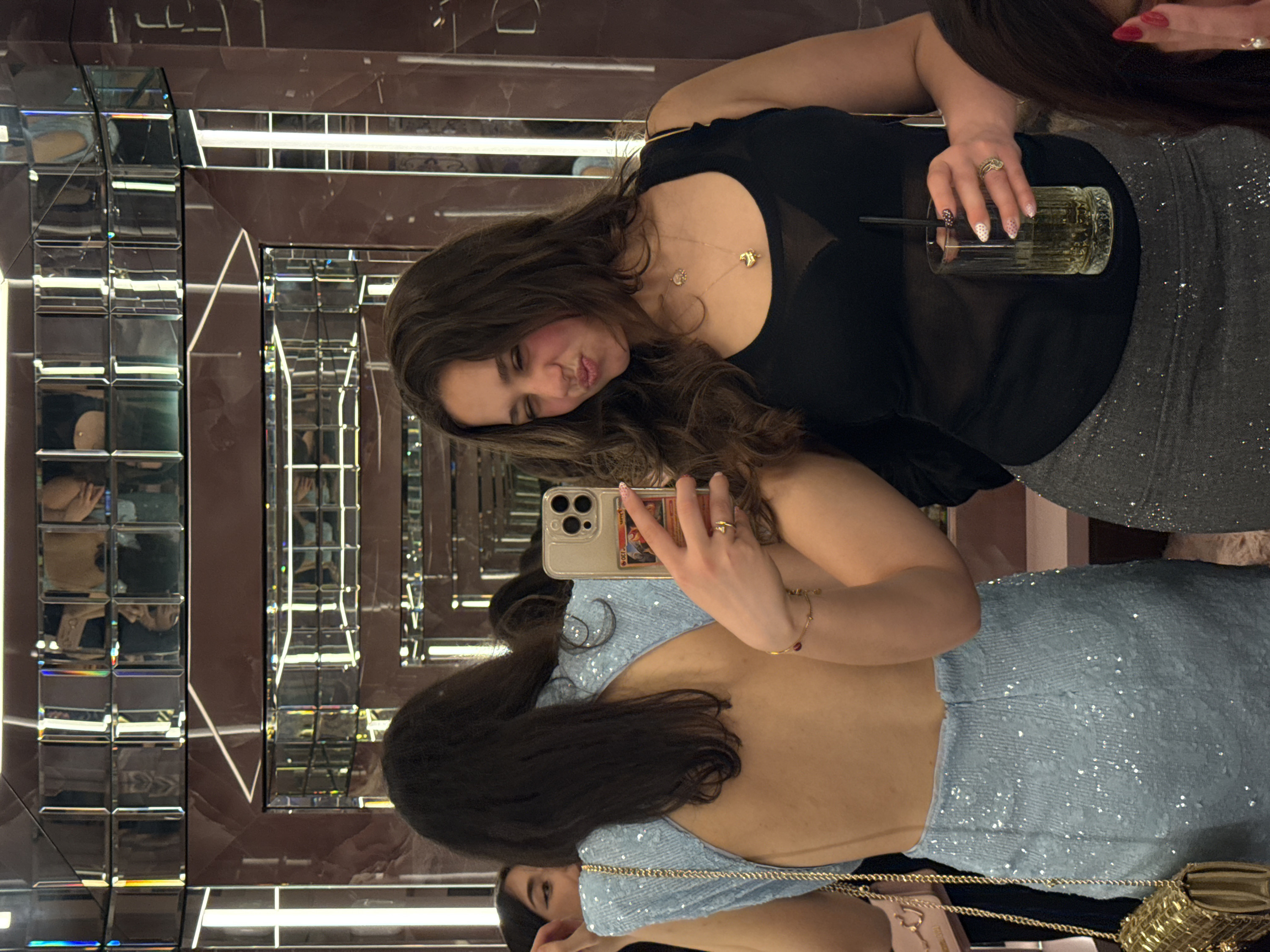The scene depicts two young adult females inside an upscale indoor establishment in Milan, Italy, likely a bar, club, or lounge. The environment features dark, polished walls extensively paneled with reflective, beveled mirrors that incorporate vertical strip lighting, creating multiple reflections and a sense of depth.
On the right, a woman with long, dark, wavy hair is taking a selfie with a light-colored smartphone held in her left hand; a trading card is visible in the phone's clear case. She makes a playful expression, winking with her right eye and puckering her lips. She wears a black sleeveless top, a sheer black bra, two gold necklaces (one with a heart pendant), a gold ring on her left ring finger, and a gold bracelet on her left wrist. Her fingernails are manicured with a white base and pink polka dots. Her right hand holds a tall, clear glass containing a dark beverage and a black straw. Her lower body is clad in dark gray, shimmering fitted bottoms.
To the left, another woman is partially visible from the back and left profile. She has long, dark hair and wears a light blue, sequined, open-back sleeveless dress. A gold chain, likely from a purse, runs over her left shoulder, and a small pink purse with a gold chain strap is visible at her left hip. Her left eye, adorned with winged eyeliner, is partially visible in the far left background. Both individuals are captured within the complex reflections of the mirrored walls.