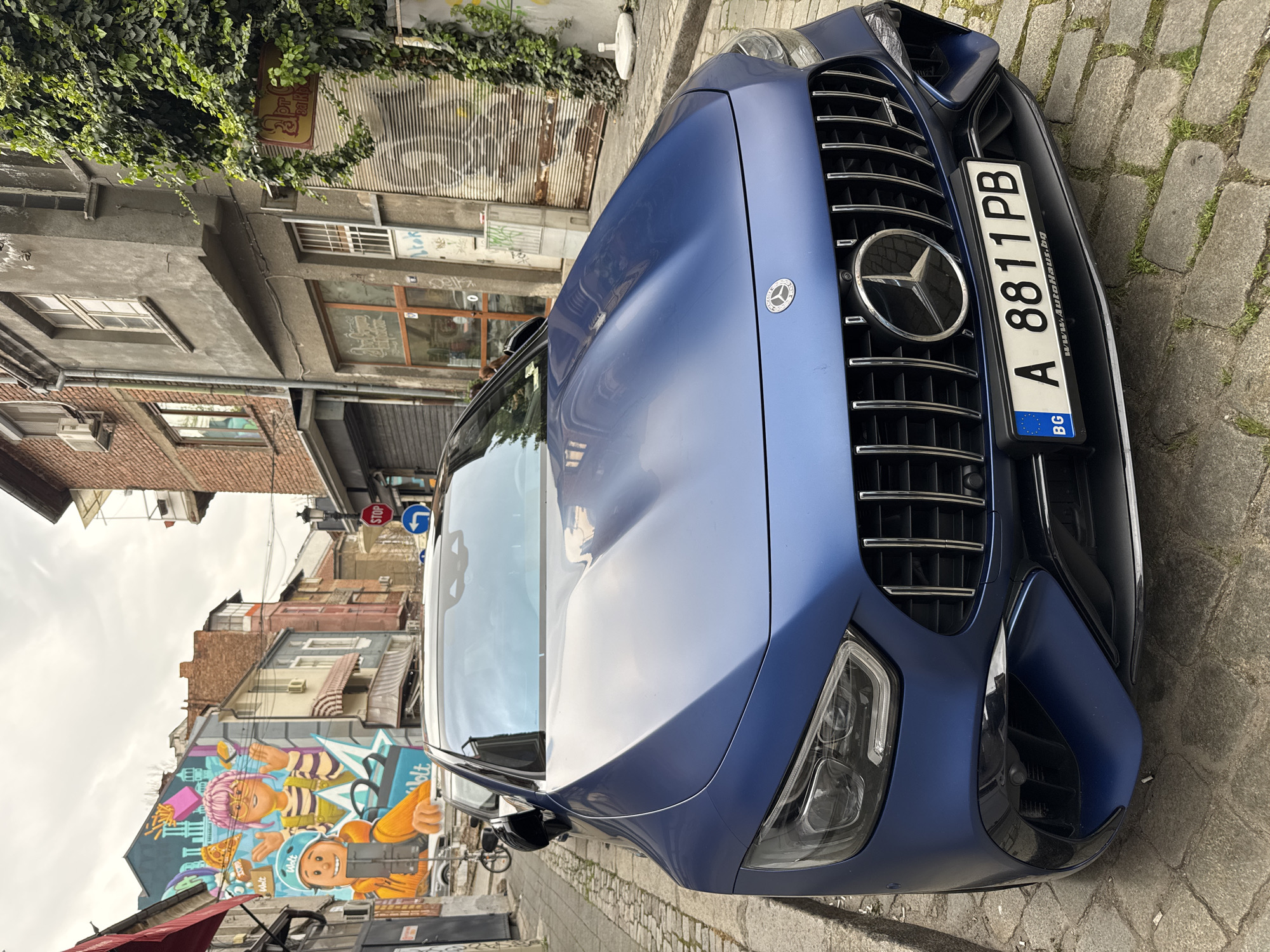 This image, taken in Plovdiv, Bulgaria, captures an urban street scene featuring a striking blue matte-finish Mercedes-Benz sports car parked on a cobblestone road. The car, prominently positioned, faces the viewer, showcasing its large grille with vertical chrome slats and the iconic Mercedes-Benz star logo. Its modern headlights are visible on either side of the front bumper. The car's license plate, mounted centrally, displays the alphanumeric code "A 8811 PB" and includes the Bulgarian country code "BG". The frame around the license plate also features the text "www.AutoHaus.bg".
The setting appears to be a narrow street or alley lined with multi-story buildings. On the left, older structures with a mix of concrete and brick facades are visible, some partially covered by green ivy. A weathered sign, mostly obscured by the foliage, might spell "Парк". On the right side, further down the street, a large and colorful mural dominates the side of a building. This vibrant graffiti art depicts two cartoonish characters: one with pink hair holding fries and pizza, and another wearing a helmet, resembling a delivery person, holding a mobile device. The brand name "Wolt" is clearly visible within the mural.
The scene appears to be set during daytime, under what seems to be an overcast sky, resulting in soft, diffused lighting. In the background, street signs are visible, including a red "STOP" sign and a blue "turn right" arrow sign, indicating an intersection. The architecture of the surrounding buildings suggests an older European city, characterized by diverse window styles, some awnings, and air conditioning units. No specific activity or people are explicitly visible; the image captures a moment of a parked car in a lively urban environment.
