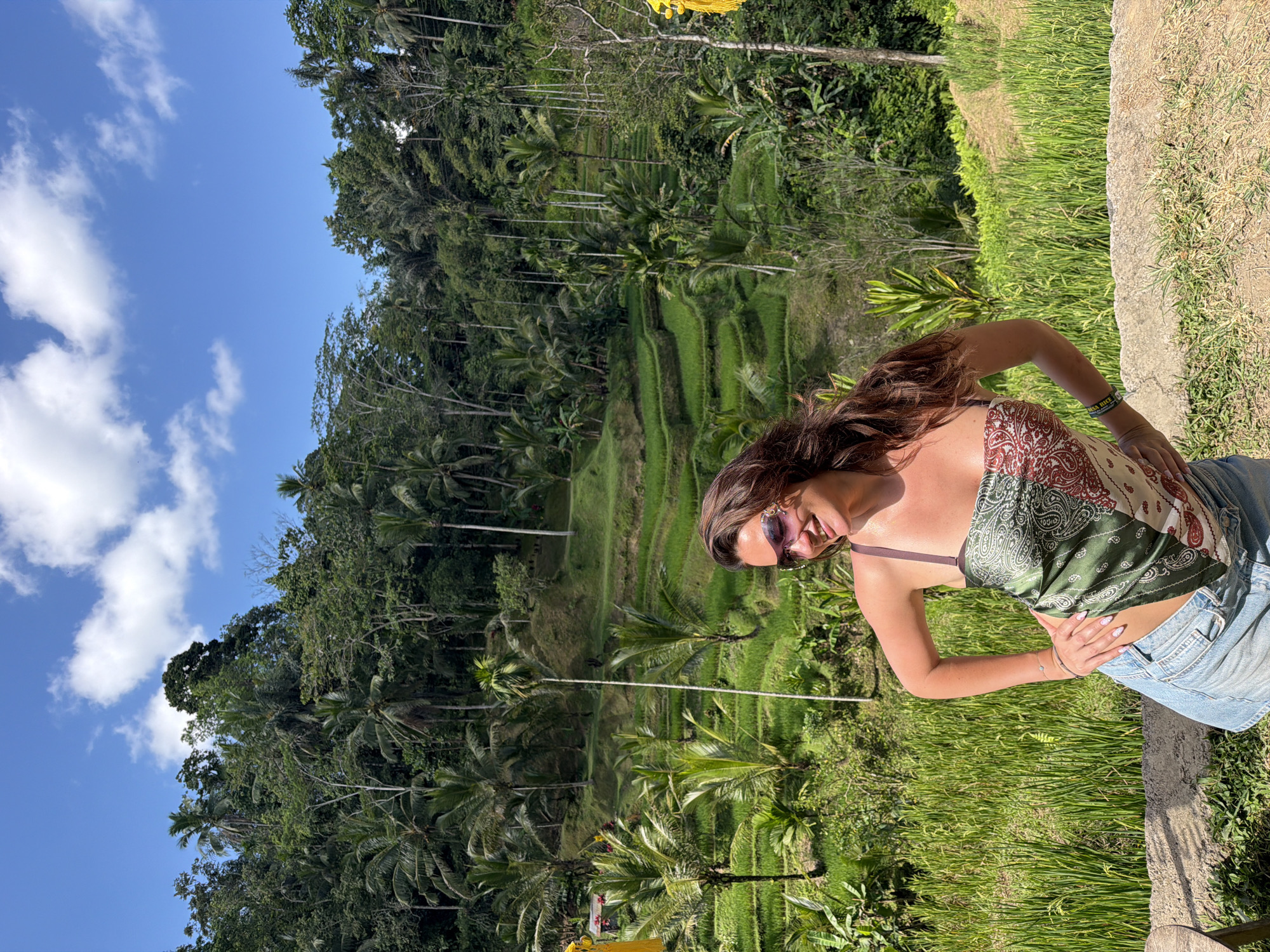A woman with long, brown, wavy hair is posed with her hands on her hips, smiling as she looks to her left. She is wearing a bandana-style top with a paisley print in shades of green, white, and red, and light-wash denim shorts. She has on ornate sunglasses with a purple tint. The background is a lush, green landscape with terraced rice paddies, palm trees, and dense jungle foliage. The sky above is blue with scattered white clouds. The scene appears to be in the Tegallalang region of Bali, Indonesia, known for its scenic rice terraces.