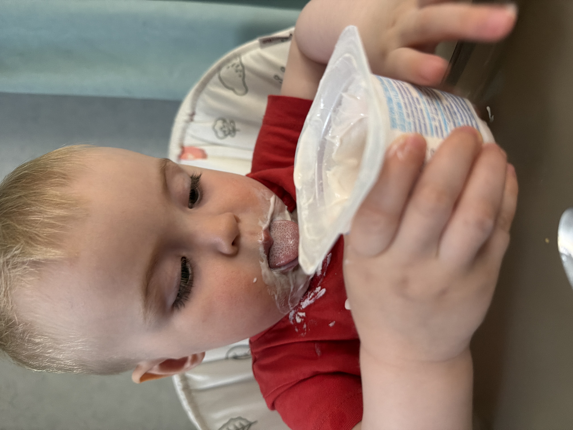 A young child is sitting in a highchair, holding a partially eaten container of yogurt. The child's face and red shirt are smeared with yogurt. The background consists of the highchair's patterned seat and a glimpse of a light-colored wall. There is no visible information to indicate the location is Votkinsk, Russia.