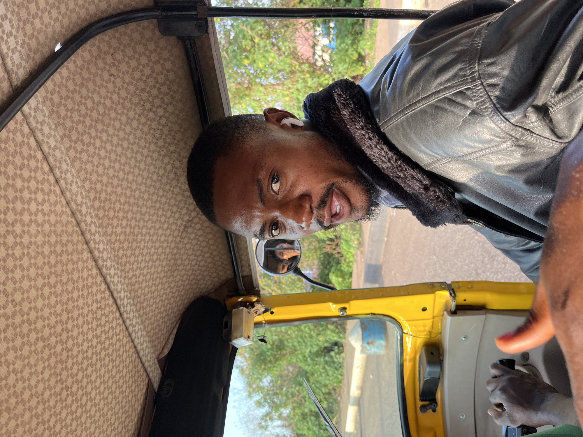The image captures the interior of a yellow three-wheeled auto-rickshaw, commonly referred to as a Keke NAPEP, in Gura Top, Nigeria. The perspective is from the front passenger position, looking forward and to the right at the driver. The driver is a dark-skinned adult male with short dark hair and a neatly trimmed beard. He is wearing a dark, possibly black, leather jacket with a contrasting dark fur or fleece collar. A white earbud is visible in his right ear. He is looking directly at the camera with his mouth slightly open. His left hand is partially visible on the vehicle's handlebar-style steering mechanism in the lower left quadrant.
In the vehicle's circular side mirror, positioned to the driver's left, the reflection of a dark-haired female is visible, likely another passenger in the vehicle. The interior roof of the Keke NAPEP features a beige fabric lining with a repeating small square or checkered pattern. Yellow metal framing and a dark structural bar are also visible inside the vehicle.
Outside the vehicle, a paved road with a light-colored curb runs alongside. The environment is rich with green vegetation, including numerous trees and bushes under a clear blue sky, indicating daytime conditions. A blue and white cylindrical object, possibly a drum, and a light-colored conical object are visible on the roadside in the background. The arm and hand of a person taking the photograph are partially visible in the lower left foreground, and a thumb is seen in the lower right foreground.