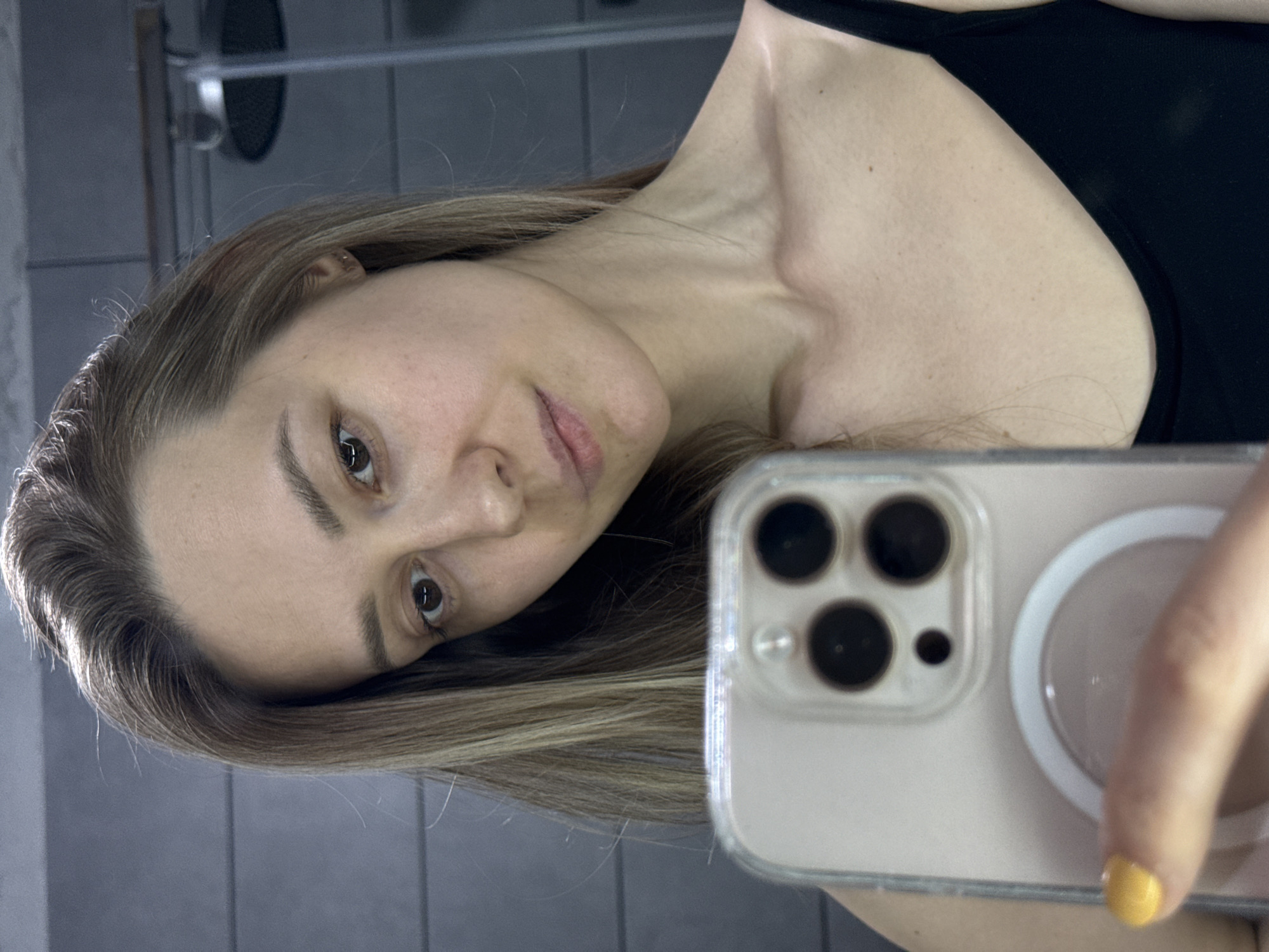 A woman is taking a selfie indoors, likely in a bathroom. She is holding a smartphone with a visible camera lens array. The woman has light brown hair and is wearing a black tank top. The background appears to be a tiled wall, potentially in a bathroom. The image does not provide information to confirm the location in Chaikovsky, Russia.