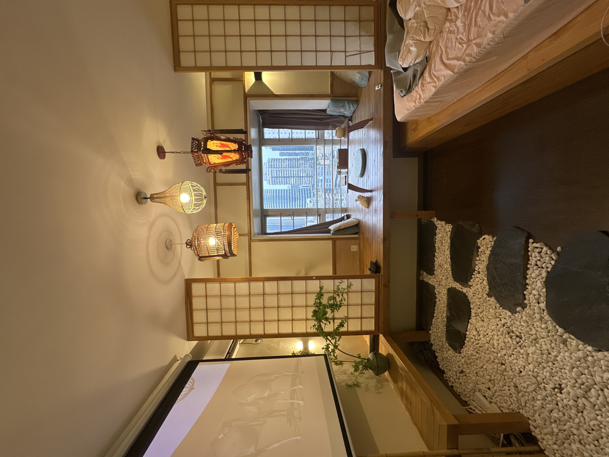 The image depicts an interior space with Japanese-inspired design elements. There's a bed with bedding, various hanging lanterns, shoji screens, and a window with a cityscape view. The space also features a decorative rock and pebble garden. The window view suggests a high-rise urban environment. While I cannot confirm the exact location within the city, the cityscape visible through the window is consistent with the architecture and density of Yuzhong District, China.