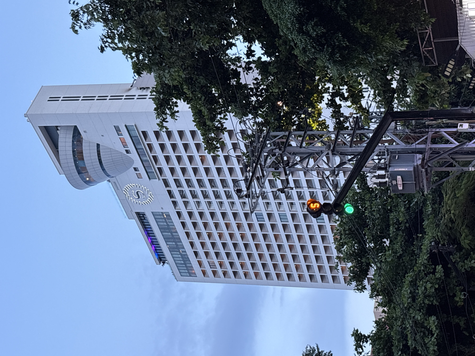 The image shows a view of the Sheraton Nha Trang Hotel & Spa located in Nha Trang Ward, Vietnam. The hotel is a tall, white building with numerous balconies. In the foreground, a traffic light displays a green light, and a portion of an electrical transformer and utility pole are visible. Lush green trees partially obscure the view of the hotel and traffic infrastructure. The sky is overcast and a pale blue.