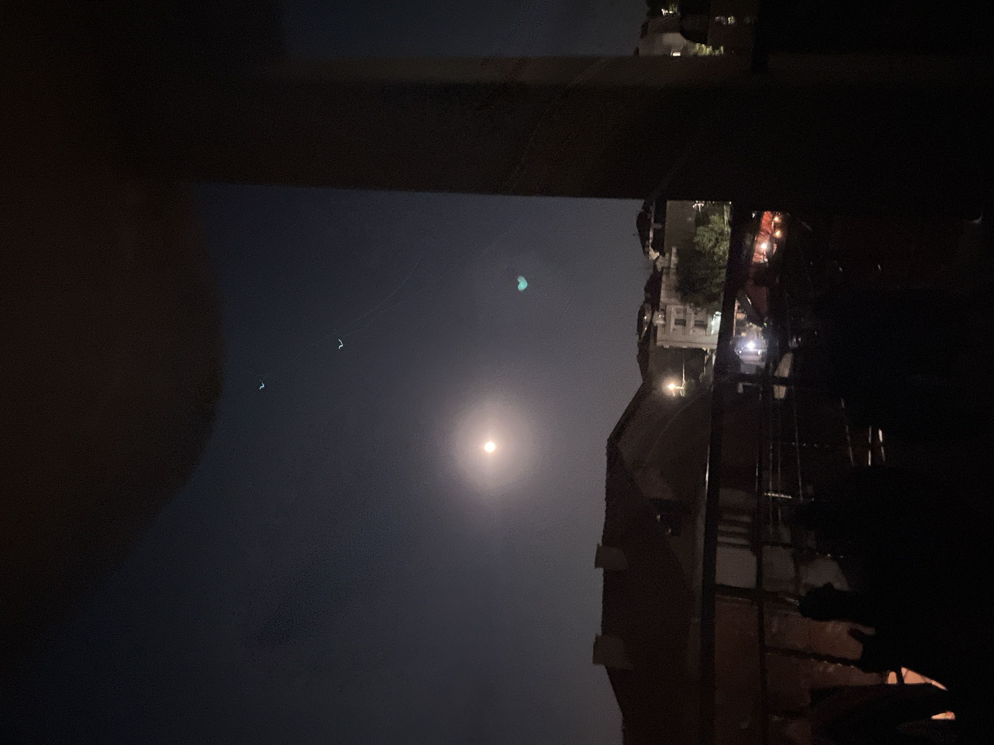The image depicts a nighttime scene over the city of Obzor, Bulgaria, as viewed from an elevated vantage point, likely a balcony or rooftop. The sky is dark, with a prominent, luminous moon creating a soft glow. Several faint, unidentifiable lights are visible in the sky, appearing as small, distant points. Below the sky, silhouetted rooftops of residential buildings are discernible, their forms softened by the dim lighting. A thick, dark vertical column, possibly a structural element of the viewing platform, dominates the right side of the frame, partially obscuring the view. In the foreground, the scene is largely shadowed, showing the indistinct shapes of what appear to be people gathered on the platform, with some indication of a railing or barrier. The overall atmosphere is one of a quiet, urban night.