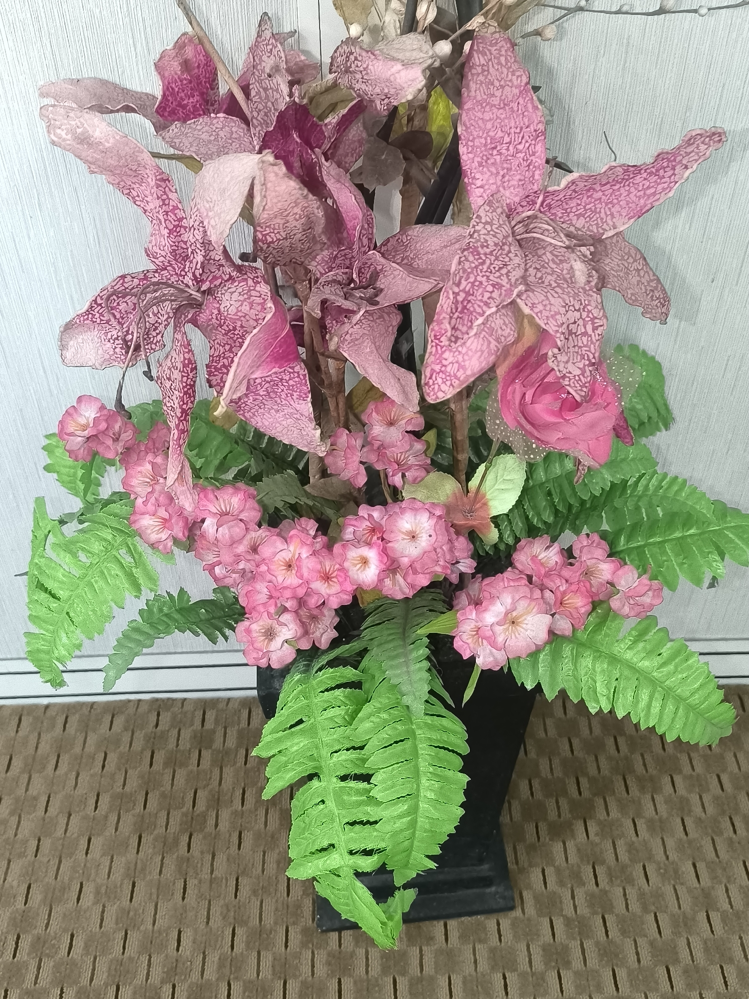 A floral arrangement of artificial pink orchids, roses, and ferns in a black vase sits on a brown textured surface, possibly a rug or mat, in front of a light-colored, textured wall. The orchids are large and prominent, with delicate pink markings on their petals. Smaller pink flowers are clustered at the base of the arrangement, interspersed with vibrant green fern fronds. The lighting is diffused, suggesting an indoor setting. The scene appears to be a static decorative display, with no people or discernible actions taking place. The overall impression is one of a carefully arranged artificial bouquet, likely for interior decoration. The location is specified as Kalabbirang, Indonesia, though there are no specific visual cues in the image to confirm this locale beyond the provided metadata.