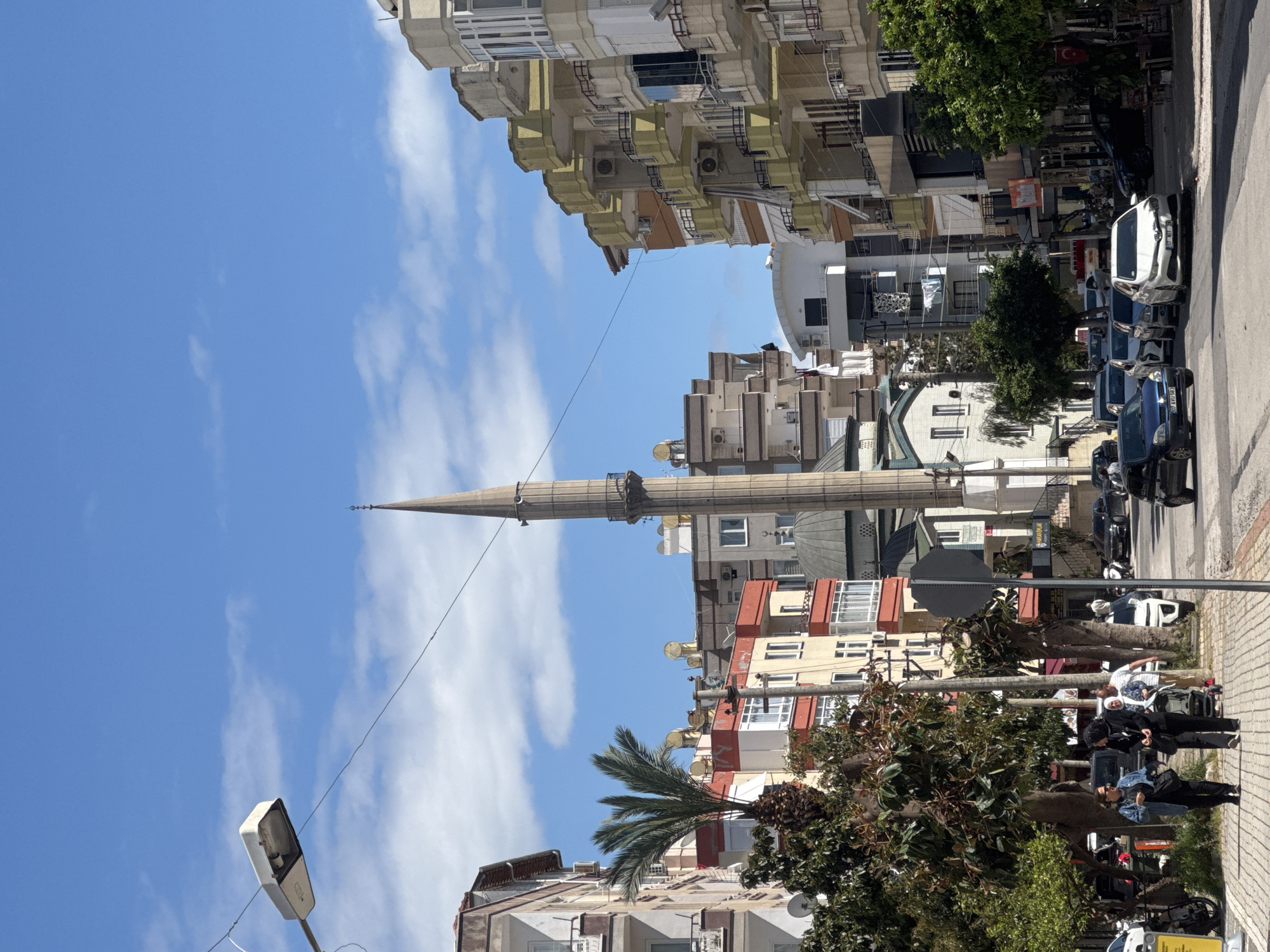 The image captures a street scene in an urban environment. A tall minaret dominates the view, indicating the presence of a mosque. Buildings of varying heights line the street, displaying a mix of architectural styles and colors. Trees and other vegetation add greenery to the scene. People are visible on the sidewalk, some standing and others sitting. Parked cars line the street. The sky is blue with scattered clouds.