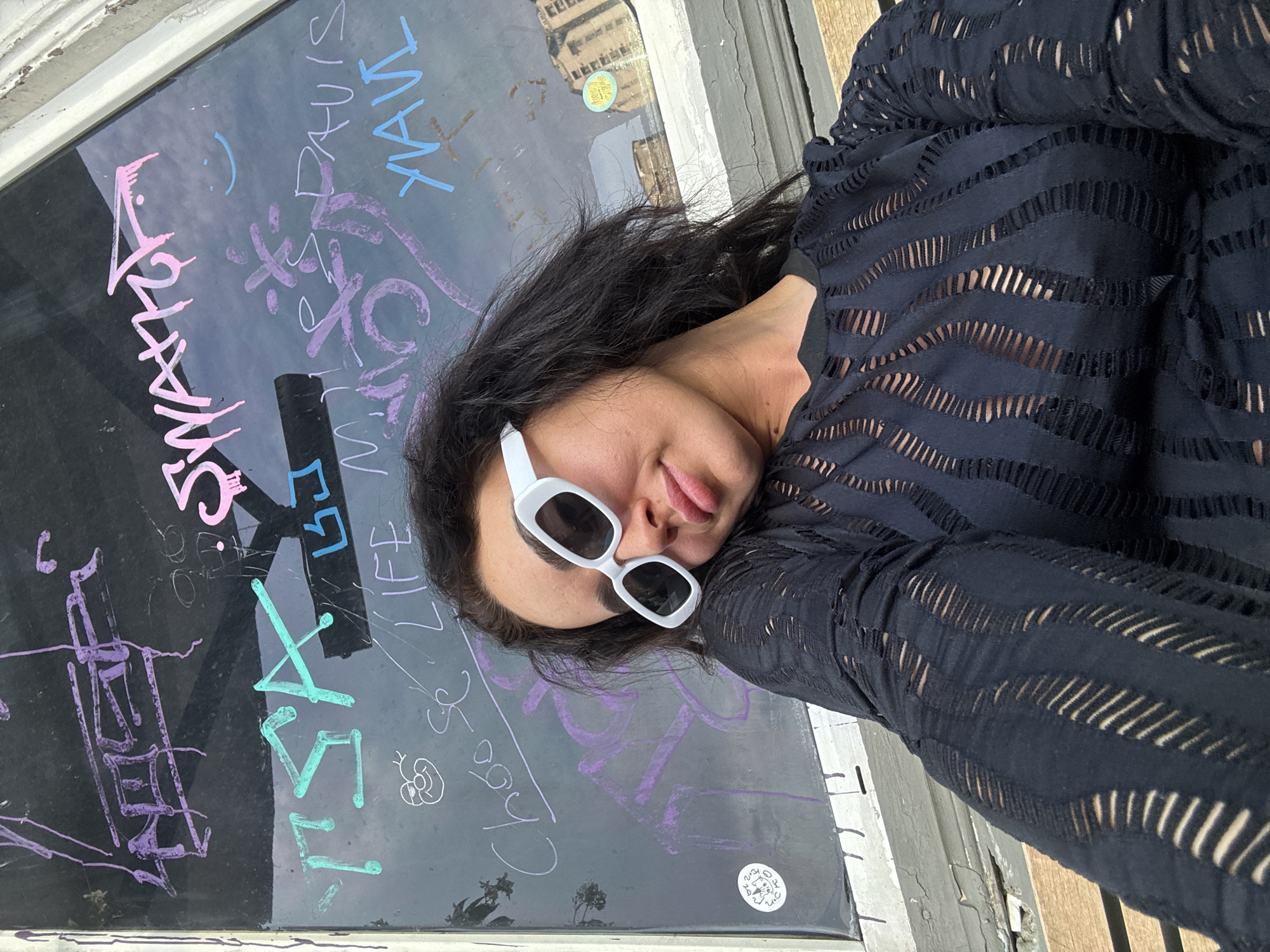 A young woman with long dark hair and white-rimmed sunglasses is taking a selfie in front of a window covered in graffiti. She is wearing a black top with cut-outs. The graffiti on the window includes words in English, such as "TSA", "LIFE", "Choose", "SWATCH", and "DAVIS". There is also a small sticker on the bottom left of the window depicting a cat. The background suggests an urban environment. The setting is the street in Tbilisi, Georgia, specifically near the Rustaveli Avenue, a main street with many shops and art galleries.