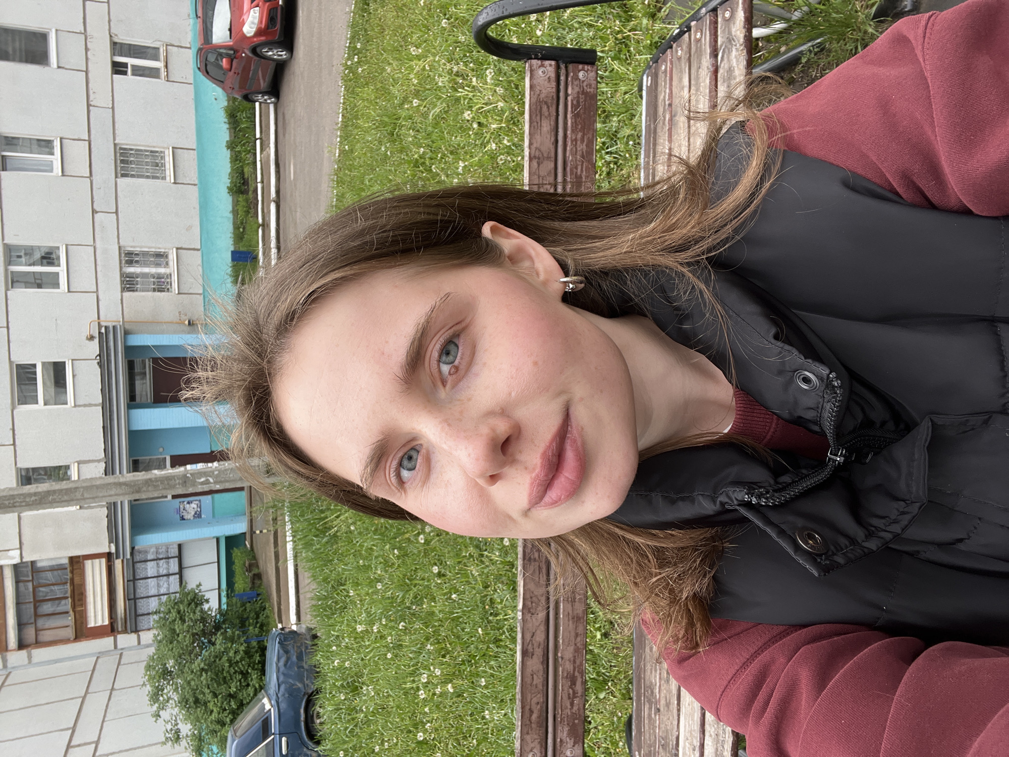 A woman is sitting on a wooden park bench in a grassy area with sparse white flowers. She is wearing a dark puffer vest over a maroon hoodie. Her hair is light brown and slightly wavy, and she has fair skin with light freckles. She is facing the camera with a slight smile.
In the background, there is a building with a light gray facade and turquoise accents around the windows and entryways. A blue car is parked near the building, partially obscured by a green tree. Another red car is visible further down the street.
The setting appears to be in a residential area of Chaikovsky, Russia, given the typical Soviet-era architecture. A concrete utility pole is present in the background.