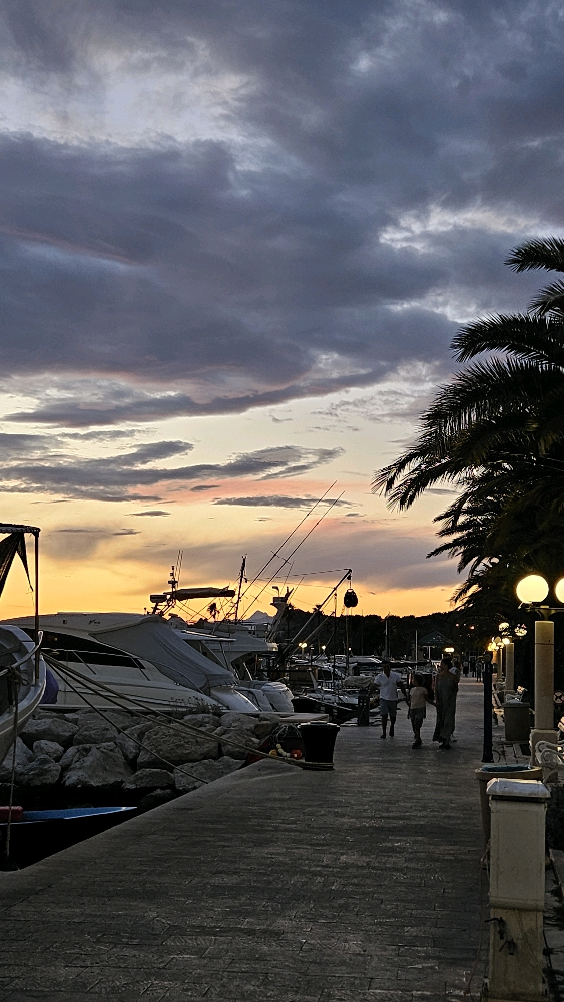 At dusk, a marina in Općina Tučepi, Croatia, is captured. The sky is painted with dramatic clouds in shades of purple, gray, and pink, contrasting with the bright orange glow of the setting sun. The foreground features a paved walkway along the water's edge, lined with lampposts that cast a warm, yellow light. To the left, several boats, including yachts, are docked and covered, resting against a rocky shore. To the right, palm trees cast shadows, and a few people are seen strolling along the walkway. The scene conveys a tranquil and serene atmosphere, typical of a coastal town at sunset.