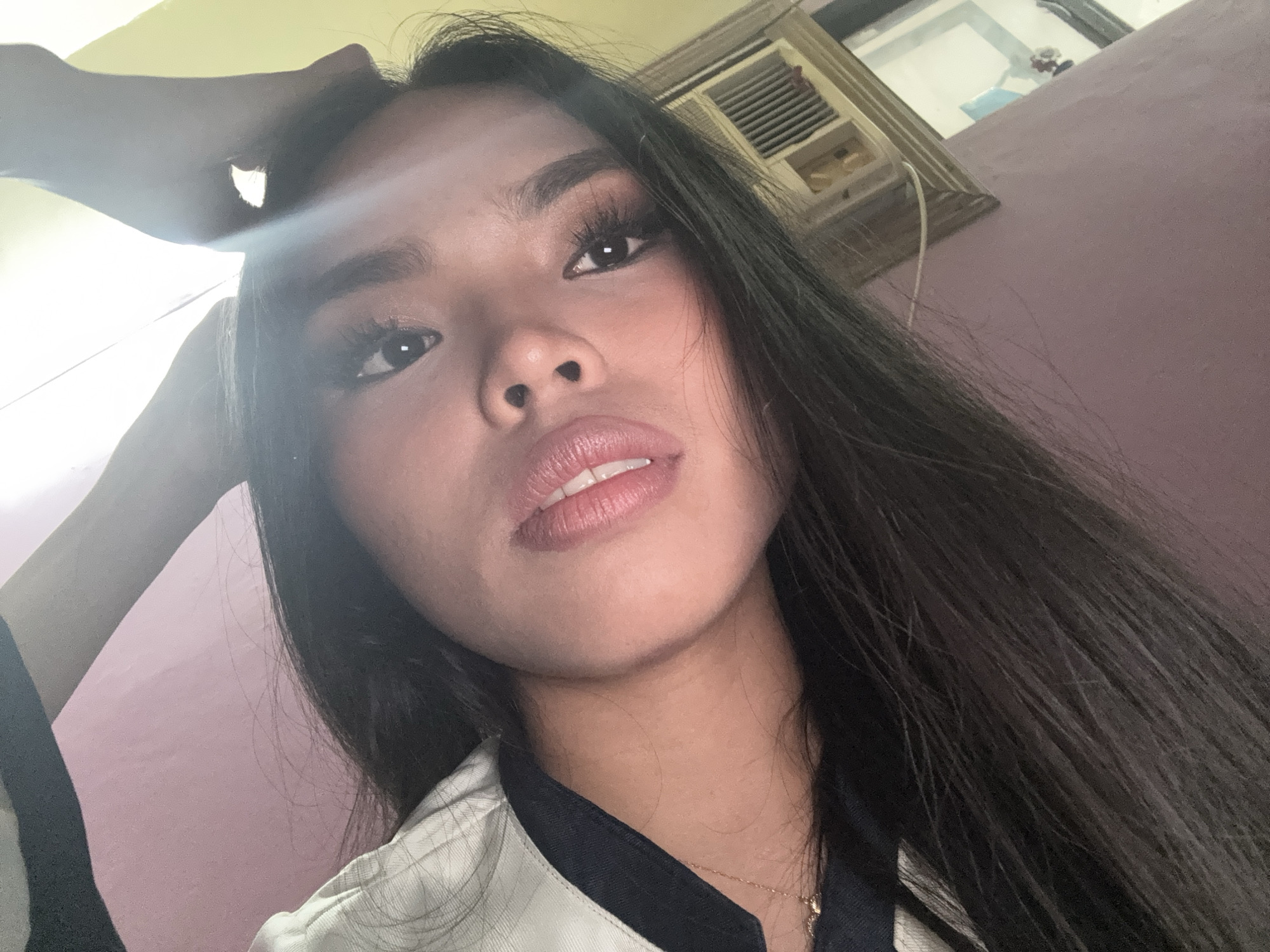 A close-up, angled selfie shows a person with long, dark hair, wearing a light-colored collared shirt over a darker garment. The person has makeup on, including mascara and lipstick. Their left hand is visible near the top of their head, with fingers extended upwards. The background appears to be an indoor setting, with a pale pink wall on the left and a yellowish wall on the right. An air conditioning unit is visible on the right side of the image. The location is not specified in the image or its context.