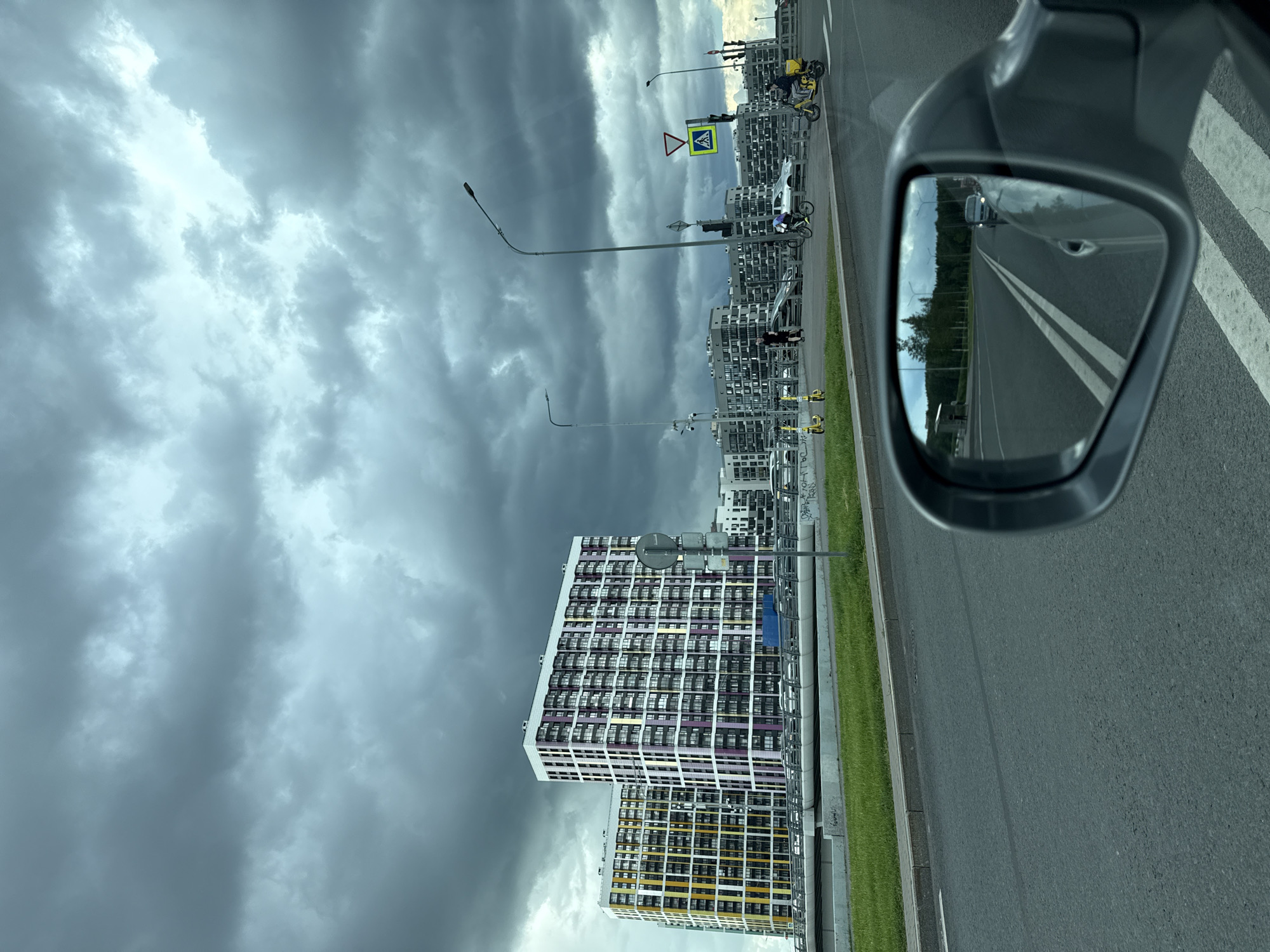 The image shows a view from a car, looking forward and slightly to the right, with the car's side mirror reflecting the road behind. The sky is overcast with dark, dramatic storm clouds. In the background, several modern, multi-story residential buildings are visible. The buildings on the left are colorful, with sections of yellow and purple. A highway overpass with a barrier runs horizontally across the mid-ground. On the overpass and the street level below, there are a few people, bicycles, and scooters. One person is riding a yellow scooter, and another is riding a bicycle. Traffic signs are visible, including a pedestrian crossing sign. The foreground consists of asphalt road with white lane markings. The reflection in the side mirror shows a road with trees and a few cars in the distance. This scene is consistent with the Nevsky Style residential complex in St. Petersburg, Russia, known for its distinctive architecture and location near transportation infrastructure.