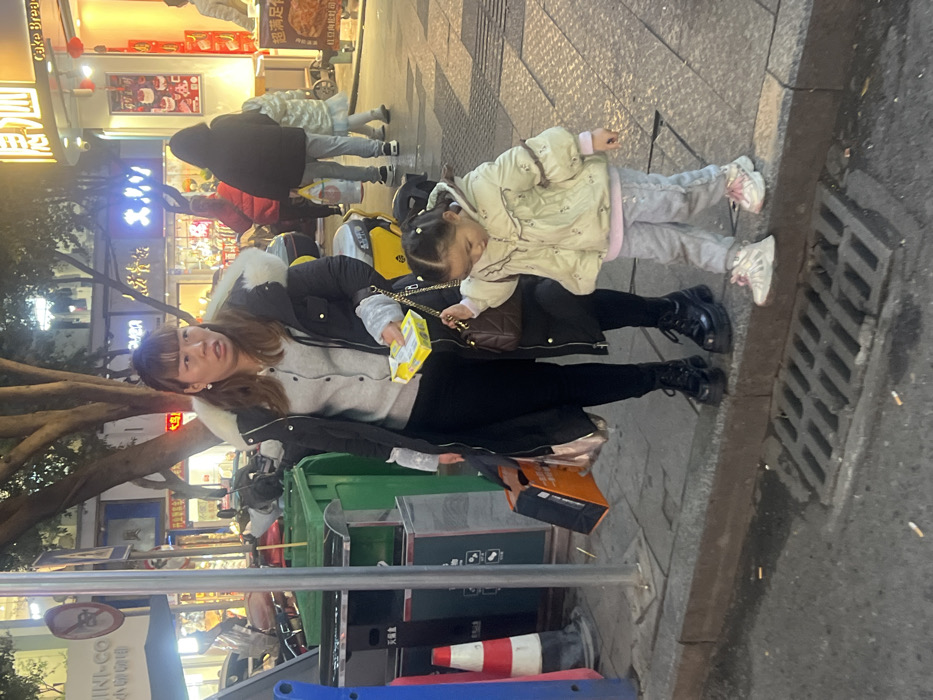 An adult East Asian female and a young East Asian female are positioned on a concrete sidewalk in an urban setting, identified as Jiangjin, China. The adult female, appearing to be in her mid-30s, possesses brown hair with bangs and wears a black winter coat with a white fur-lined hood, a light gray cardigan, black pants, and black platform boots. She holds a small yellow rectangular packet in her left hand, a dark brown handbag with a gold chain strap in her right hand, and two shopping bags (one black, one metallic brown/pink) are suspended from her left arm. Her expression is characterized by wide eyes, directed towards the right side of the frame.
The young female, estimated to be between 3 and 5 years old, has her hair styled in two small pigtails. She wears a light cream-colored quilted puffer jacket with ruffled details, a light purple long-sleeved shirt, light gray pants with subtle embellishments, and light-colored athletic shoes with pink laces. She holds the chain strap of the adult female's handbag with her left hand and is in the action of stepping from the asphalt street onto the sidewalk curb with her right foot, looking downwards.
The immediate environment includes a metal storm drain grate embedded in the asphalt street, with several discarded cigarette butts visible nearby. On the sidewalk to the left, two green public trash receptacles are present, alongside a partially visible red and white striped traffic cone and a metal pole. The background features multiple illuminated storefronts with Chinese signage, including "MIMI-CO 小包包咖啡" (MIMI-CO Small Bag Coffee), "恩泗" (Ensi) with "Cake Bread" beneath it, and "超满足 炸鸡" (Super Satisfying Fried Chicken). Several motorcycles or scooters are parked along the sidewalk behind the trash cans. Distant trees with bare branches are visible. The scene is lit by a combination of natural daylight and artificial light from the commercial establishments.
