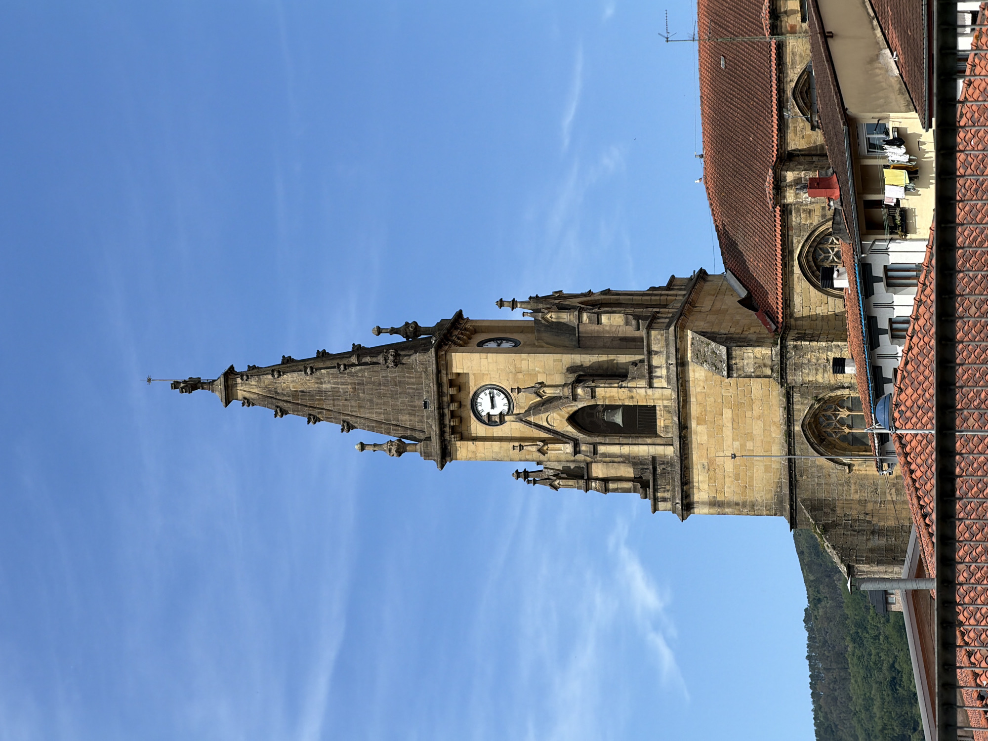 The image shows a church tower rising against a clear blue sky. The tower is made of light-colored stone and features a clock face. The style is Gothic, with spires and decorative elements. To the right of the tower are buildings with red-tiled roofs, suggesting the church is in a densely built urban area. Laundry is hanging on a line from one of the buildings. In the background, a treed hill is visible. The location of the church is Errenteria, Spain.