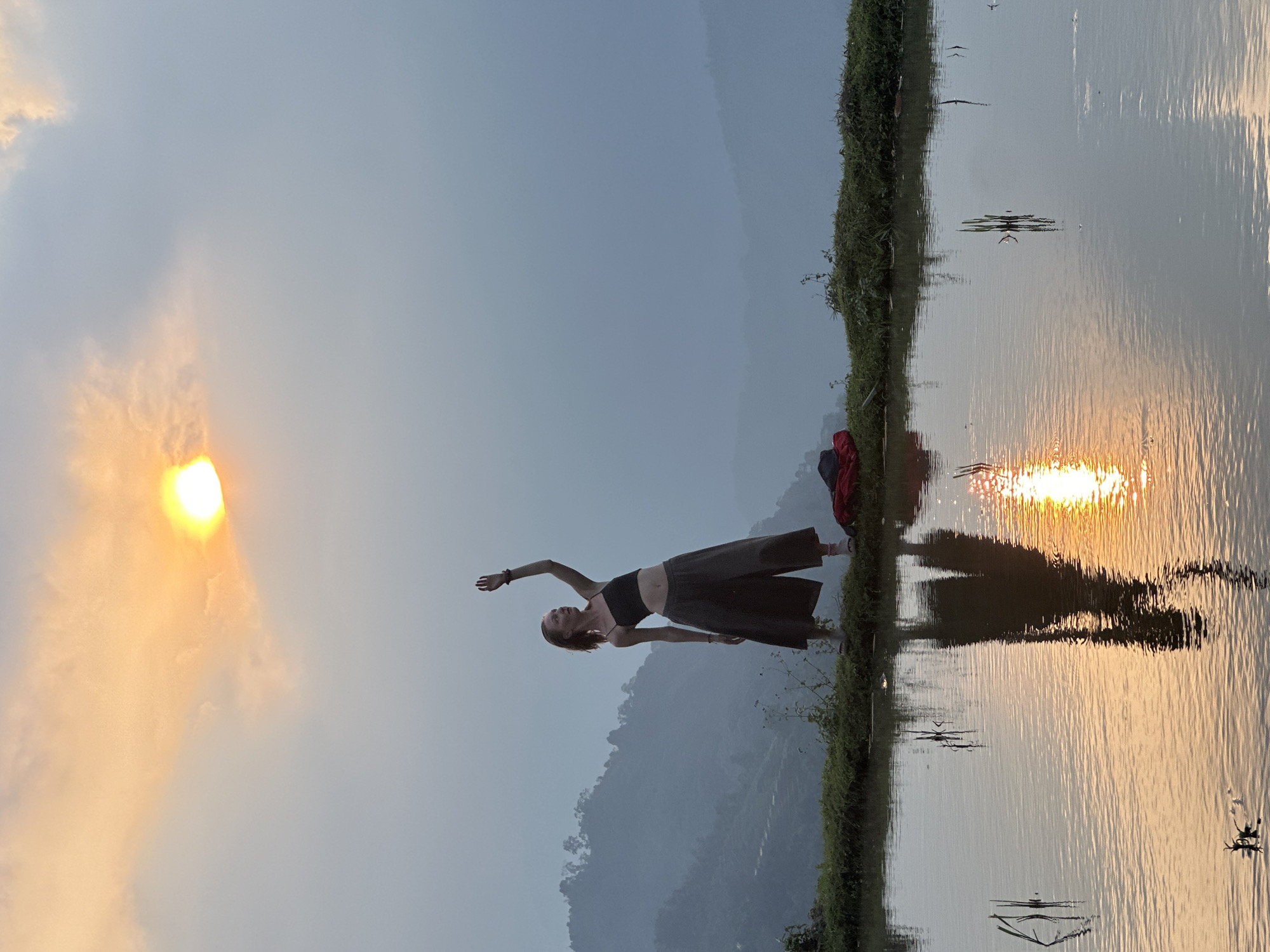 The image depicts a serene outdoor scene, likely at sunrise or sunset, set in a rural landscape, consistent with Nậm Dịch Commune, Vietnam. A woman stands on a narrow, grassy embankment that separates a calm body of water on the right from a landmass on the left.
The woman, appearing to be in an active or yoga-like pose, is the central subject. She wears a dark crop top and wide-leg pants, with her arms gracefully extended and her body slightly angled. Near her feet on the embankment, a red item, possibly a bag or folded cloth, is visible.
The sky on the left side of the frame features a bright, radiant sun, partially obscured by soft, illuminated clouds, casting a warm, golden light. This golden light is vividly reflected on the rippled surface of the water, creating a prominent streak of orange and yellow that extends towards the bottom right. Beyond the woman, in the midground, a range of mountains or hills rises, their contours softened by atmospheric haze. The tranquil water, the reflective surface, and the expansive sky with the setting or rising sun contribute to an overall atmosphere of peace and natural beauty. No text is visible in the image.
