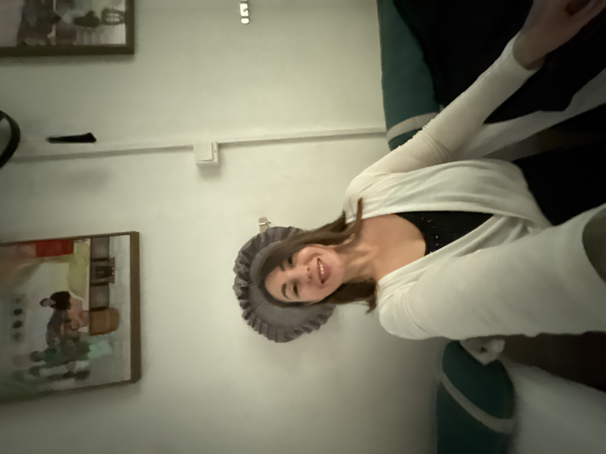 A woman wearing a grey ruffled hat and a white cardigan over a black top poses for a selfie in a room in Huizhou, China. She is smiling and looking at the camera. The background includes a wall with two framed pictures, a light switch, and part of a couch with green and white striped cushions. The overall lighting is dim, casting shadows on the wall.