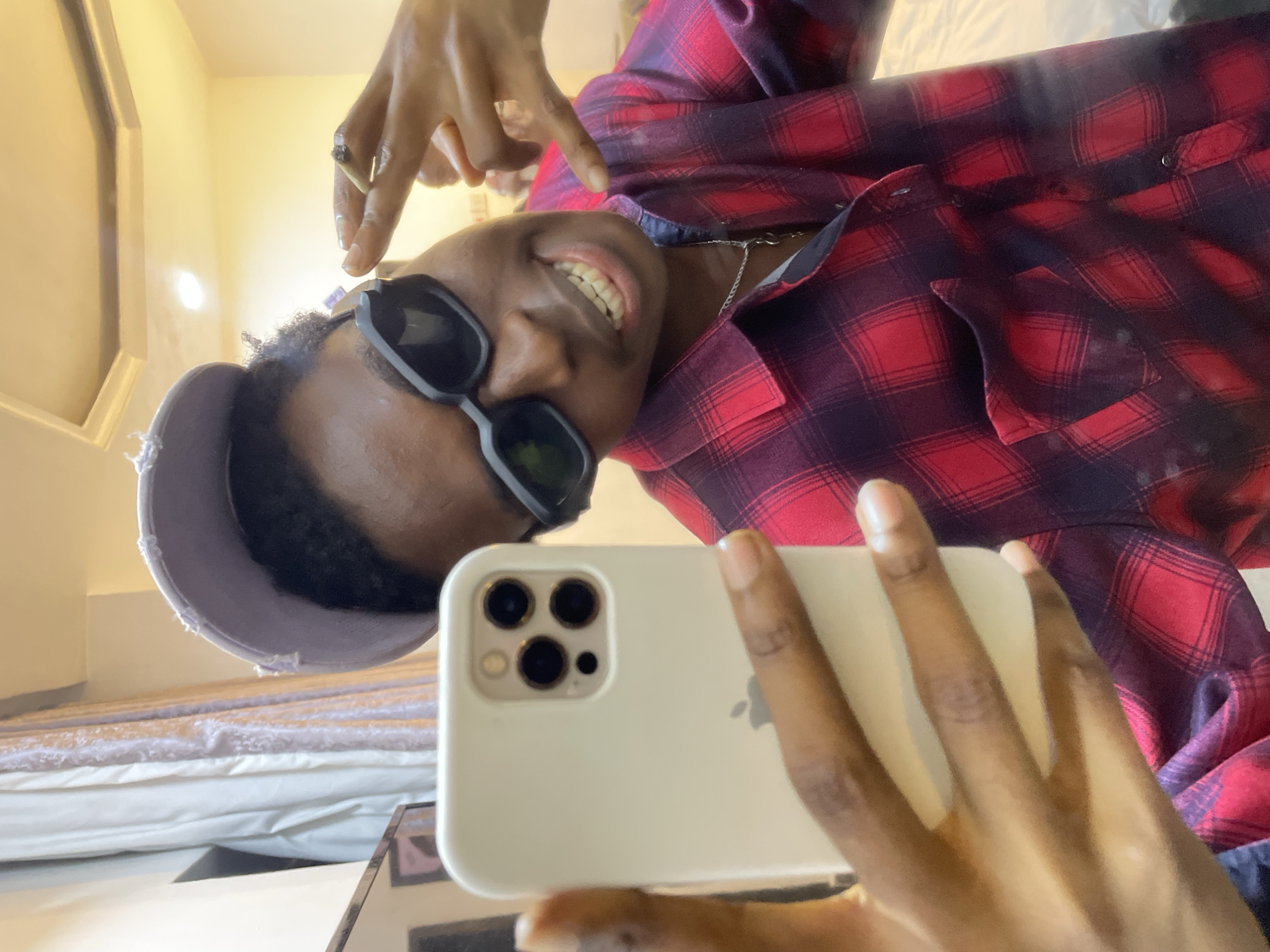 The image depicts a young person taking a selfie, possibly in a room. The person has dark skin, is wearing a red and black plaid shirt, sunglasses, and a gray cap. They are smiling, and their teeth are visible. One hand is holding a white iPhone with a triple-lens camera, and the other hand is raised, holding a lit joint. The background shows a wall and a bed with light-colored bedding. The location is stated as Awka, Nigeria.