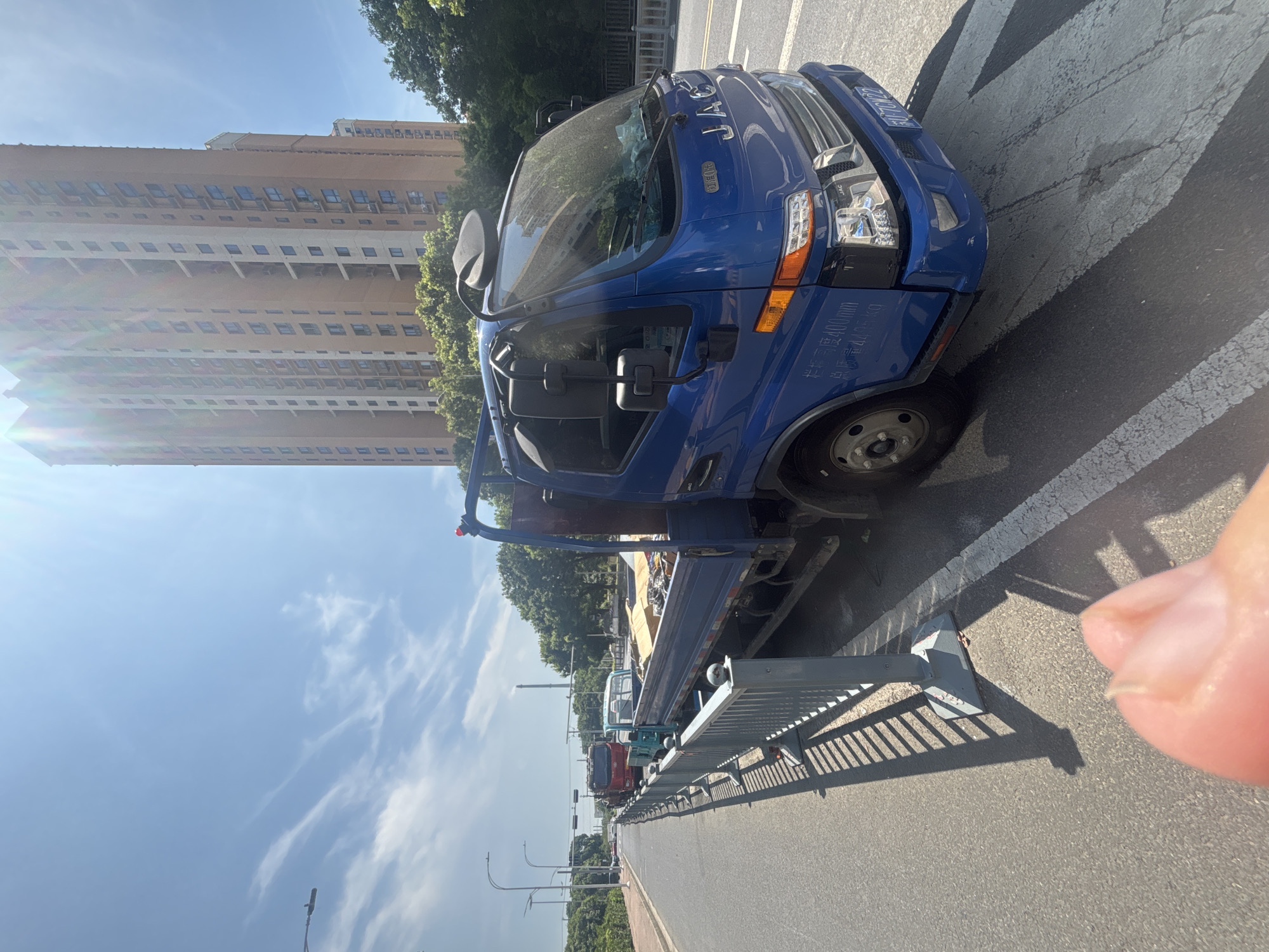A bright, sunny day in Suzhou Industrial Park, China, with a blue JAC truck parked on the side of the road. The truck is loaded with debris. In the background, a tall residential building stands under a clear blue sky with wispy clouds. Other vehicles and trees are visible along the street. A person's thumb is in the foreground, out of focus.