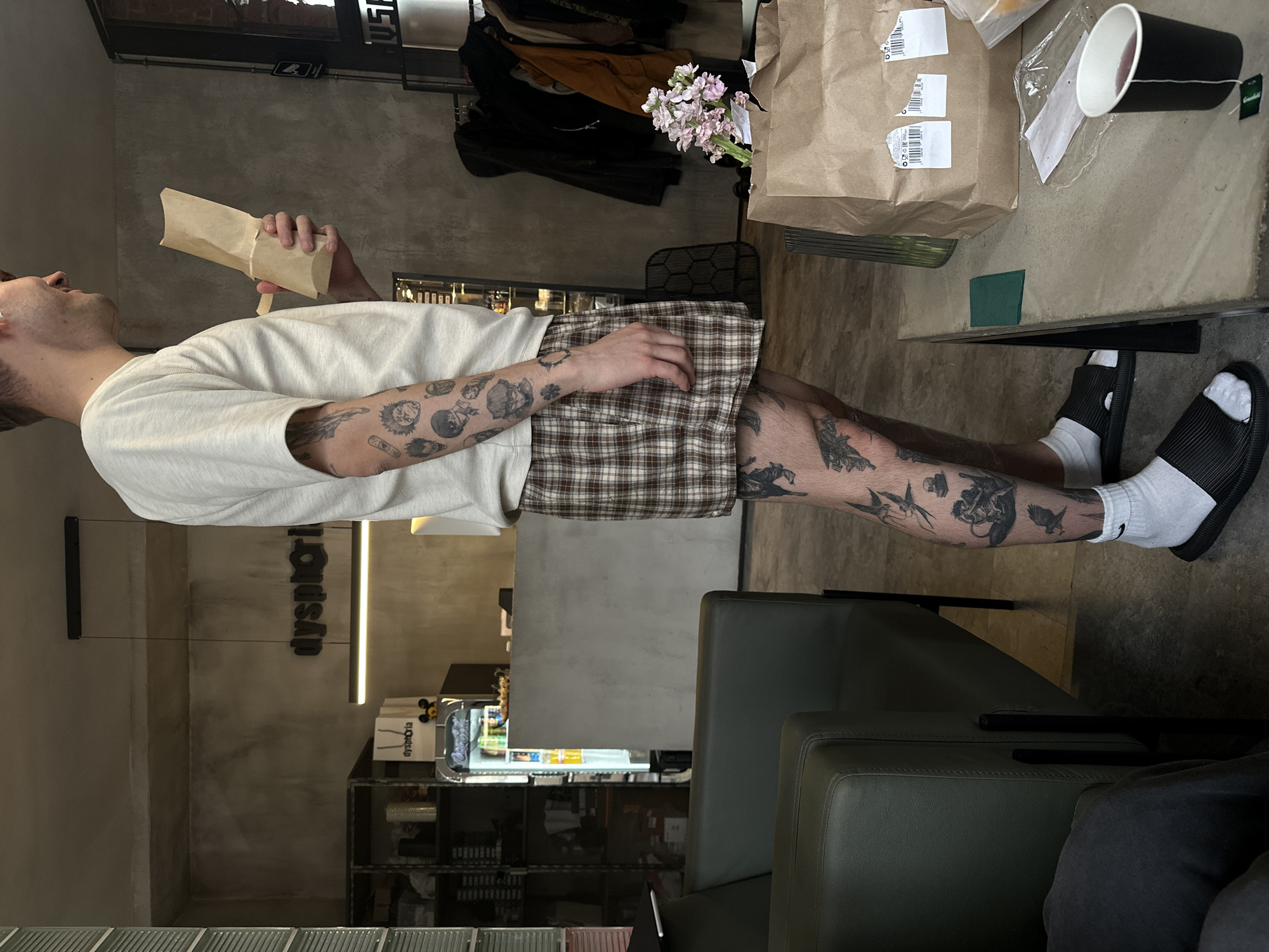 A man stands at a table in a brightly lit interior space, likely a cafe, restaurant, or business, holding a brown paper bag. He wears a white t-shirt, plaid shorts, white socks, and black sandals, and has tattoos on his arms and legs. The table has a brown paper bag, disposable cup, and other items on it. A leather chair is visible beside the table. The background reveals a shelf stocked with goods, and a sign that reads "dysphoria" is visible on the wall. The location is likely in Moscow, Russia.