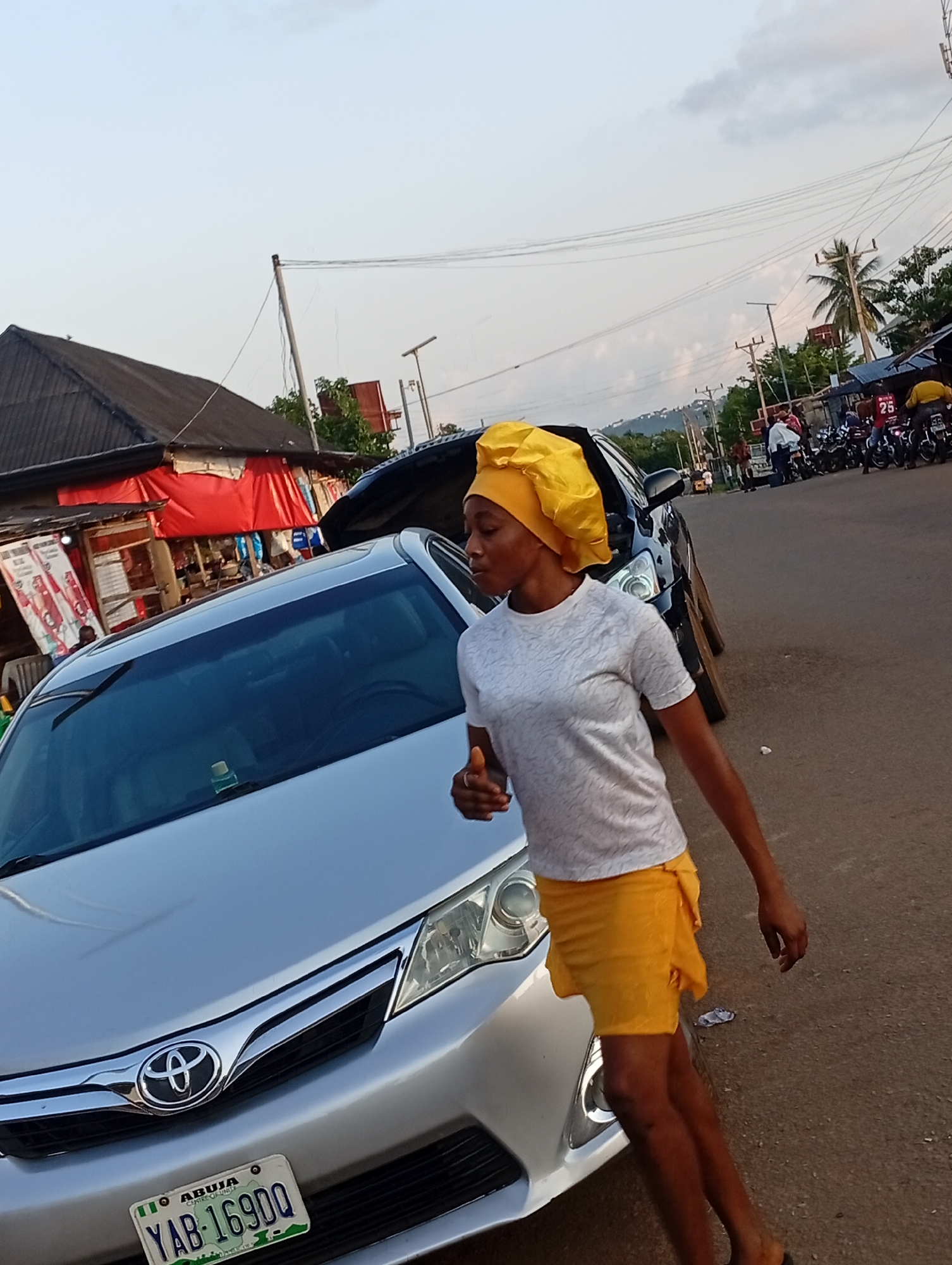 A young woman wearing a yellow headscarf and a white patterned t-shirt with yellow shorts is standing next to a silver Toyota Camry. The car has a license plate that reads "ABUJA YAB-16900". The woman is looking to her left and has her right hand raised. She appears to be in motion. In the background, there are several motorcycles parked along the side of a paved road. A small market with red tarps and stalls is visible to the left. Overhead, power lines crisscross the sky. The scene is set in Nsugbe, Nigeria.