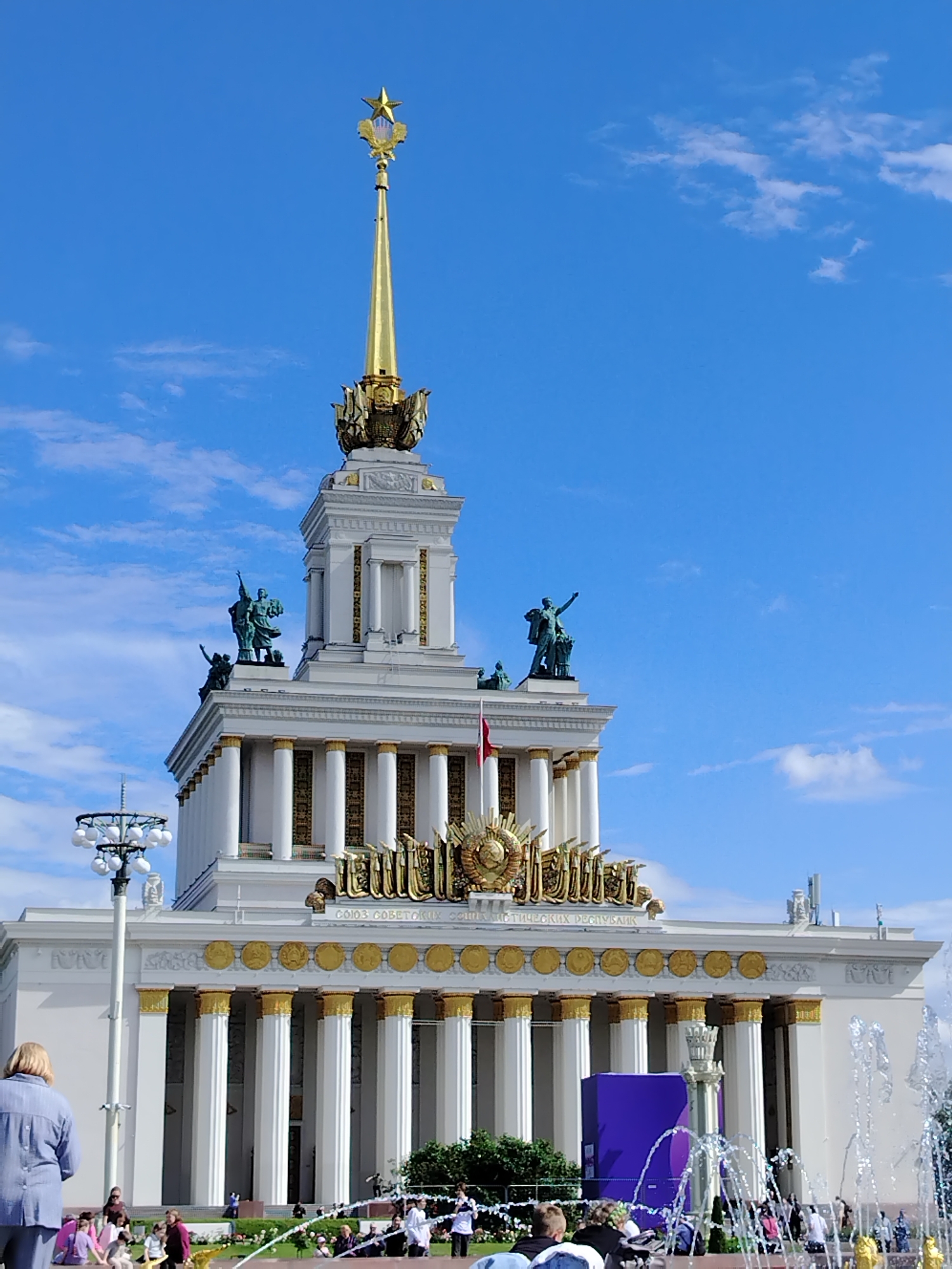 The image depicts the iconic Pavilion "Friendship of Peoples" at VDNKh (Exhibition of Achievements of National Economy) in Moscow, Russia. The ornate pavilion, a prominent example of Stalinist architecture, is bathed in sunlight under a clear blue sky with scattered white clouds. Its white facade is adorned with golden columns and decorative elements, including statues of figures, and a prominent golden spire topped with a star.
In the foreground, a woman in a blue shirt is standing on the left, facing away from the camera. To her right, a group of people are gathered, some sitting and others standing. A fountain is active on the right side of the frame, with jets of water spraying upwards. The overall atmosphere suggests a sunny day at a public gathering place, with visitors enjoying the architecture and recreational facilities.