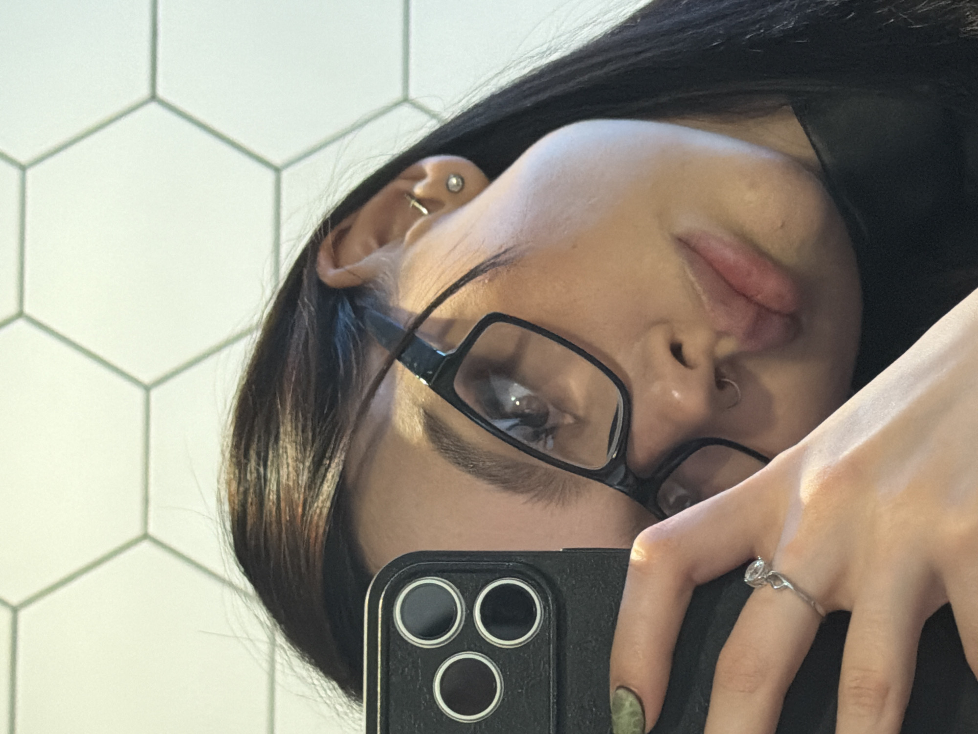 A selfie of a person with dark hair and glasses, taken at an angle, showing the right side of their face. The person has a nose piercing, ear piercings, and is wearing a ring on their ring finger. The background is a white tile wall. The person is holding a black smartphone with three camera lenses. The location is unspecified, in an unknown city.