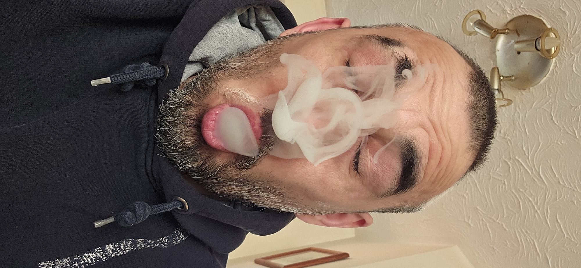 The image displays an adult male in an indoor setting, rotated 90 degrees clockwise, so his head is on the left side of the frame.
**Subjects:** The primary subject is a man with a short, greying beard and dark eyebrows. He is wearing a dark blue hooded sweatshirt with a grey lining. His eyes are closed or heavily squinted.
**Objects:** Visible objects include the man's dark blue hoodie, its drawstring with metal aglets, a wall-mounted light fixture with gold-colored accents in the upper right background (as oriented), and a small, empty wooden picture frame resting on a surface in the lower left background.
**Scene/Setting:** The scene is indoors, against a light-colored, textured wall, possibly featuring anaglypta wallpaper.
**Activity or event taking place:** The man is exhaling a dense cloud of white vapor or smoke. A distinct, elongated "bubble" or ring of vapor is emerging from his mouth, while additional vapor swirls around his face, partially obscuring his nose and forehead. This appears to be a deliberate act of exhaling vapor, possibly from a vaping device, creating a specific shape or effect.
**Notable details:** The image's orientation shows the man's head tilted to the left. The lighting is artificial, indicating an indoor environment without apparent natural light, so the time of day is not discernible. The man's facial expression suggests concentration or a physical response to the vapor. The background texture adds a subtle detail to the room. The location context provided is Norwich, United Kingdom, though no specific visual elements within the image directly confirm this.
**Visible text:** There is no visible text in the media file.