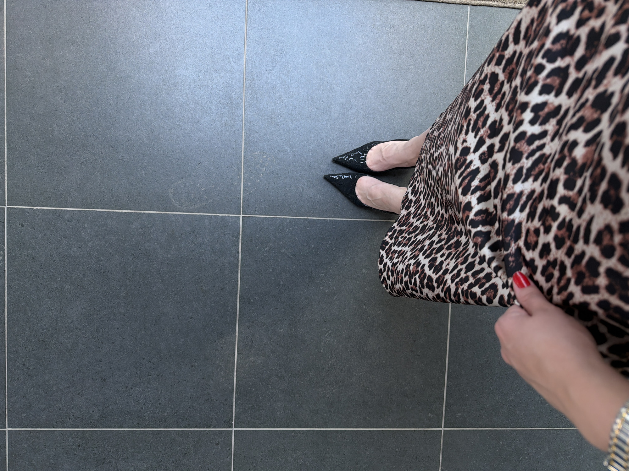 A person is standing on a grey tiled floor, wearing black pointed-toe shoes and a leopard print dress. The person's hand is visible, holding the dress. No information about the location being Izhevsk, Russia is present in the provided image.