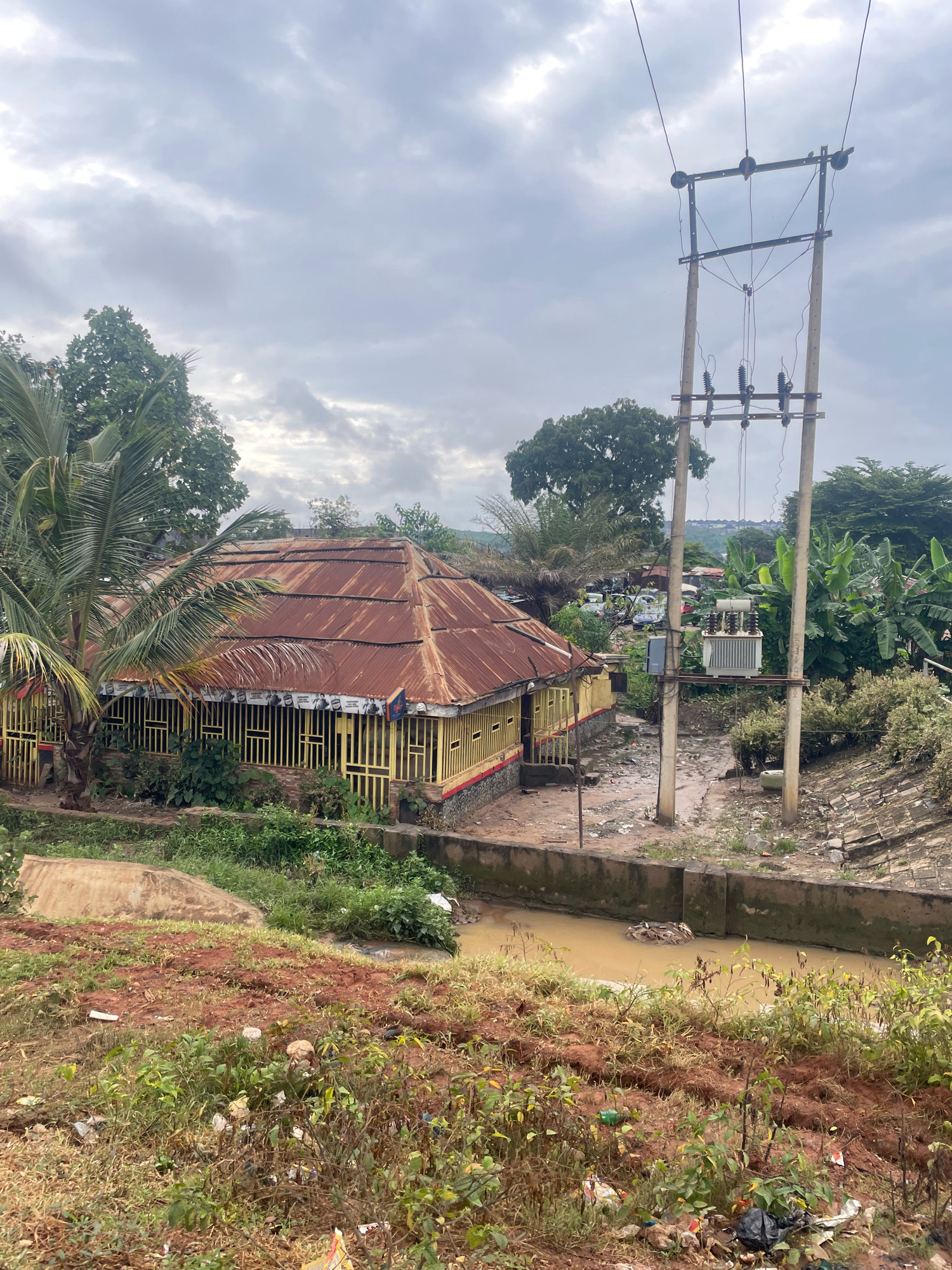 This is a mid-angle, eye-level, outdoor, daylight, full shot of a somewhat neglected urban scene in Abuja, Nigeria. The focus is on a weathered, yellow-painted building with a rusty corrugated metal roof, surrounded by dense green vegetation and palm trees. A large electrical transformer on a pole stands to the right of the building, connected by overhead wires. In the foreground, a muddy, brown stream flows, with debris and trash scattered along its banks and the surrounding red-brown earth. The sky is overcast with heavy, gray clouds, suggesting a humid or rainy climate. In the background, more trees and buildings are visible, indicating a densely populated area. The scene evokes a sense of an underdeveloped or poorly maintained urban environment.