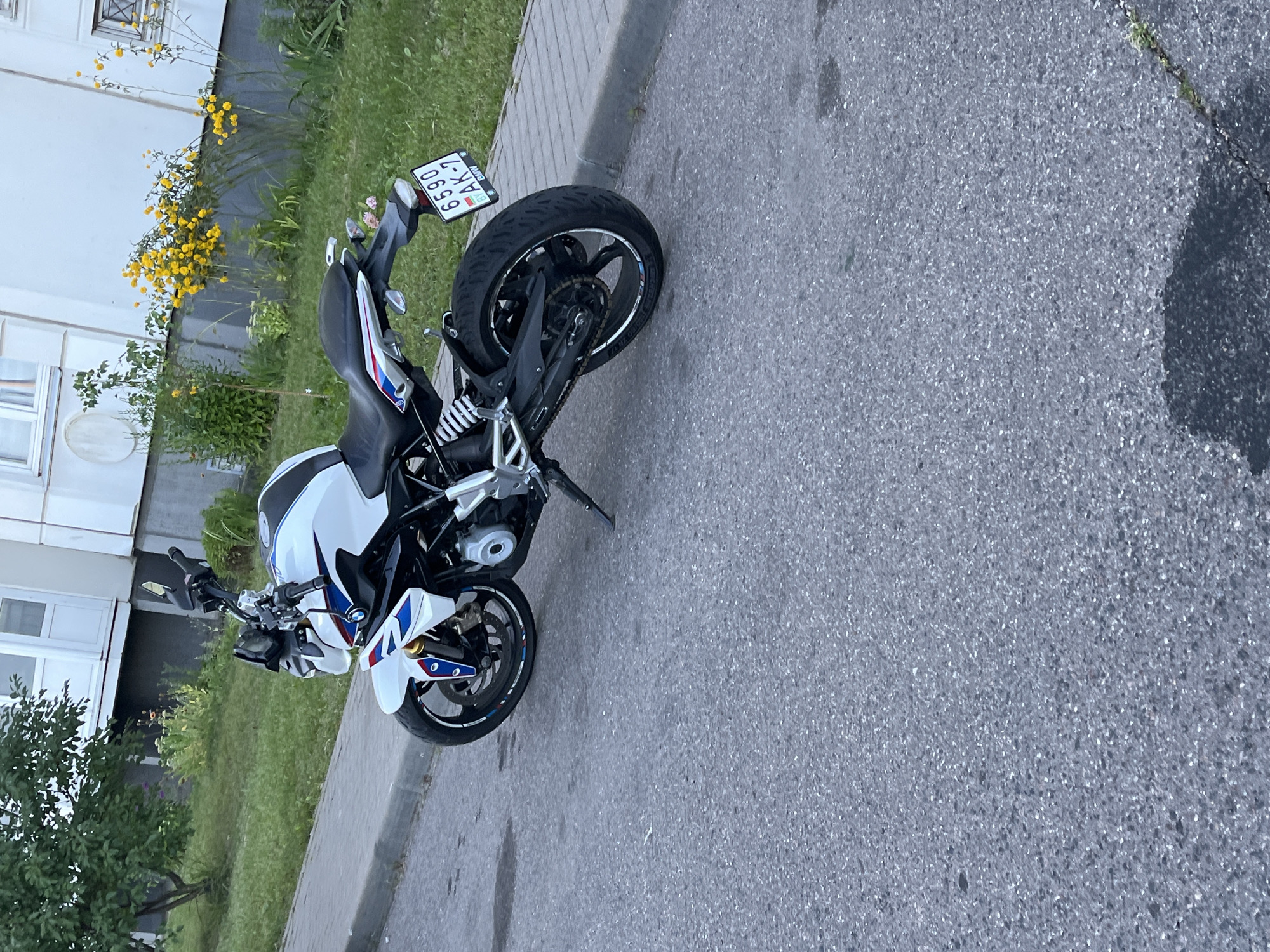 An unmounted white BMW motorcycle with blue and red accents is parked on an asphalt street next to a paved sidewalk. The motorcycle faces to the left and its license plate, reading "6590 AK-7", is visible. In the background, a building with light-colored walls and windows is visible, with green grass and yellow flowers growing in front of it. The scene appears to be a residential area in Minsk, Belarus.