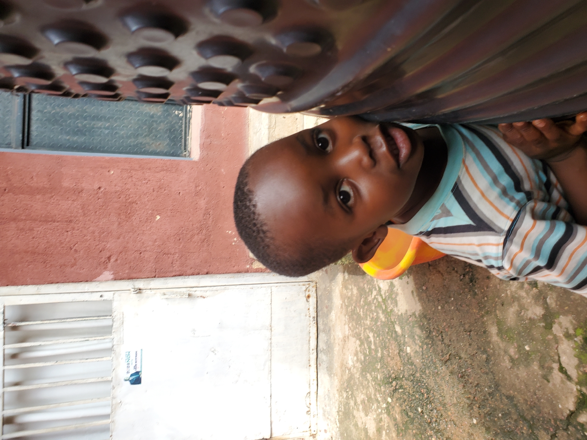 A young boy peeks out from under a dark, textured surface, possibly the underside of a table or structure, looking upwards and to his left. He has dark skin, short-cropped hair, and dark eyes. He is wearing a striped t-shirt with alternating horizontal bands of light blue, white, grey, and orange. His mouth is slightly open, revealing his teeth. A yellow and orange plastic object is visible behind his head.
The background suggests an outdoor setting with a reddish-brown wall on the left. Below this wall is a white door or panel with horizontal bars or slats. A small blue and white sticker or label is attached to the white surface. The ground appears to be a rough, uneven surface, possibly concrete or dirt, with some patches of green moss or vegetation. The lighting suggests it is daytime, with shadows indicating the sun is present. The image's orientation is rotated.